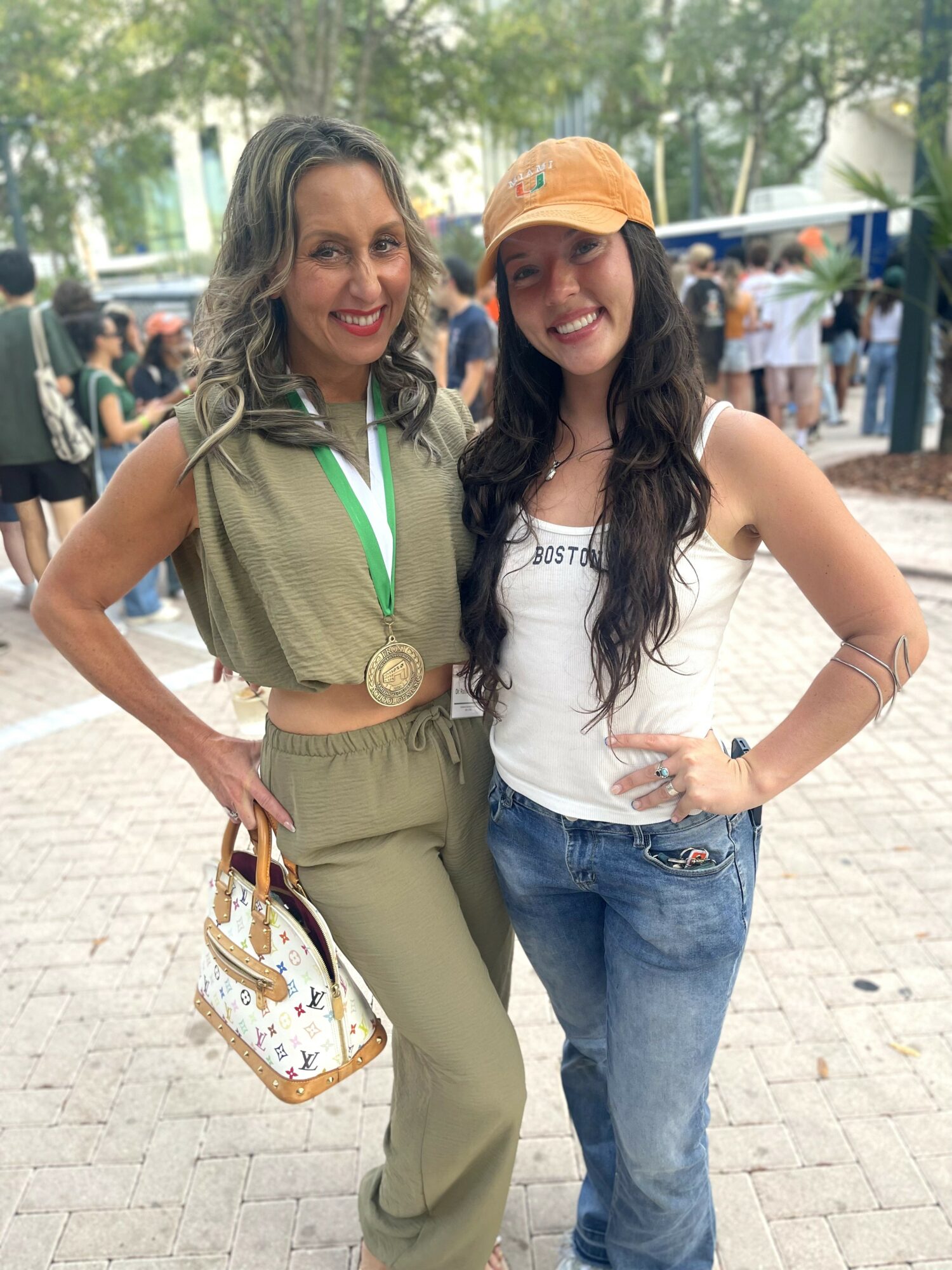 Two women smiling outdoors, one holding a handbag, surrounded by people and trees.