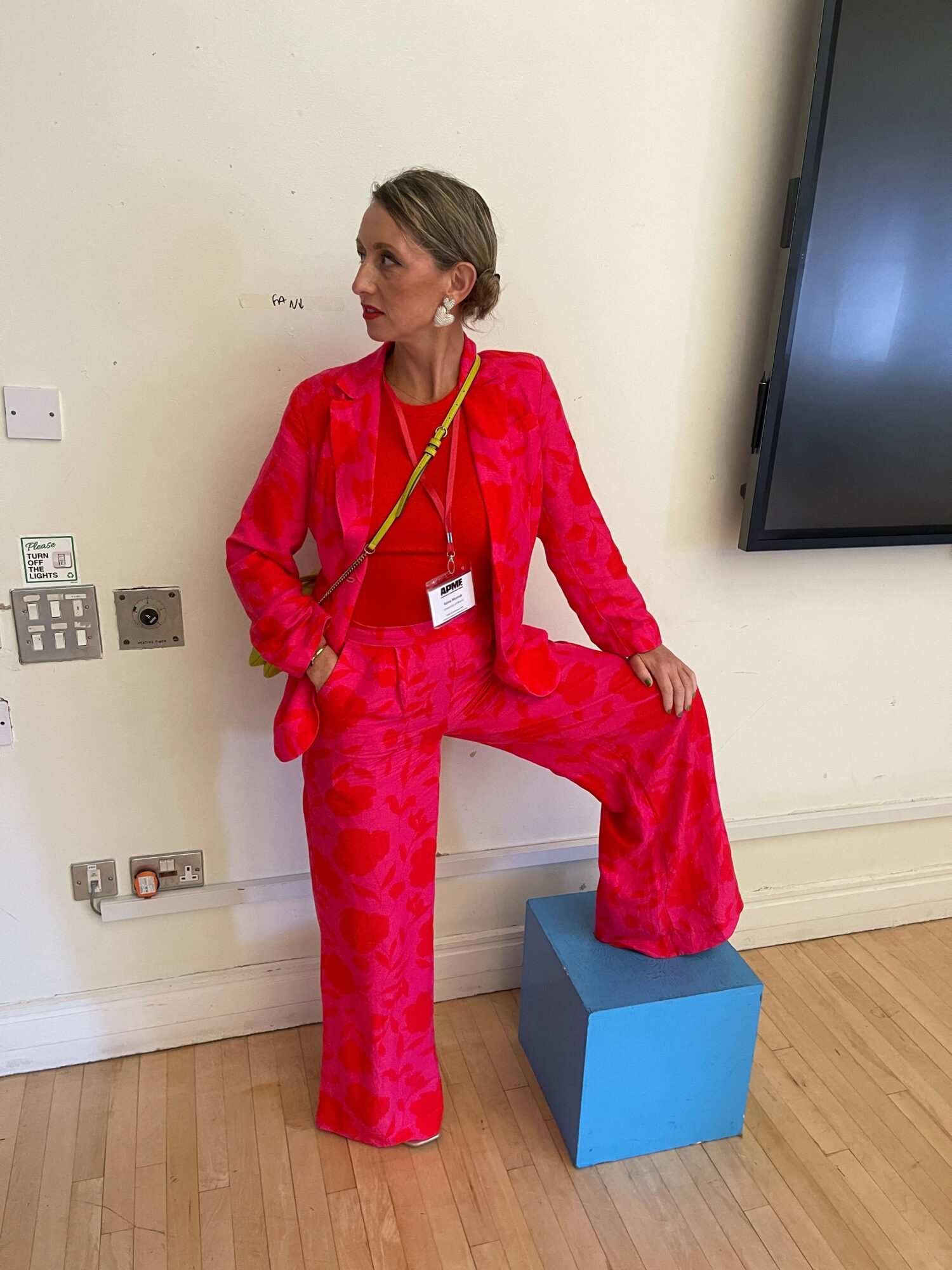 Person in red outfit with a yellow measuring tape around neck, standing next to a blue cube, in a room with wooden floor.