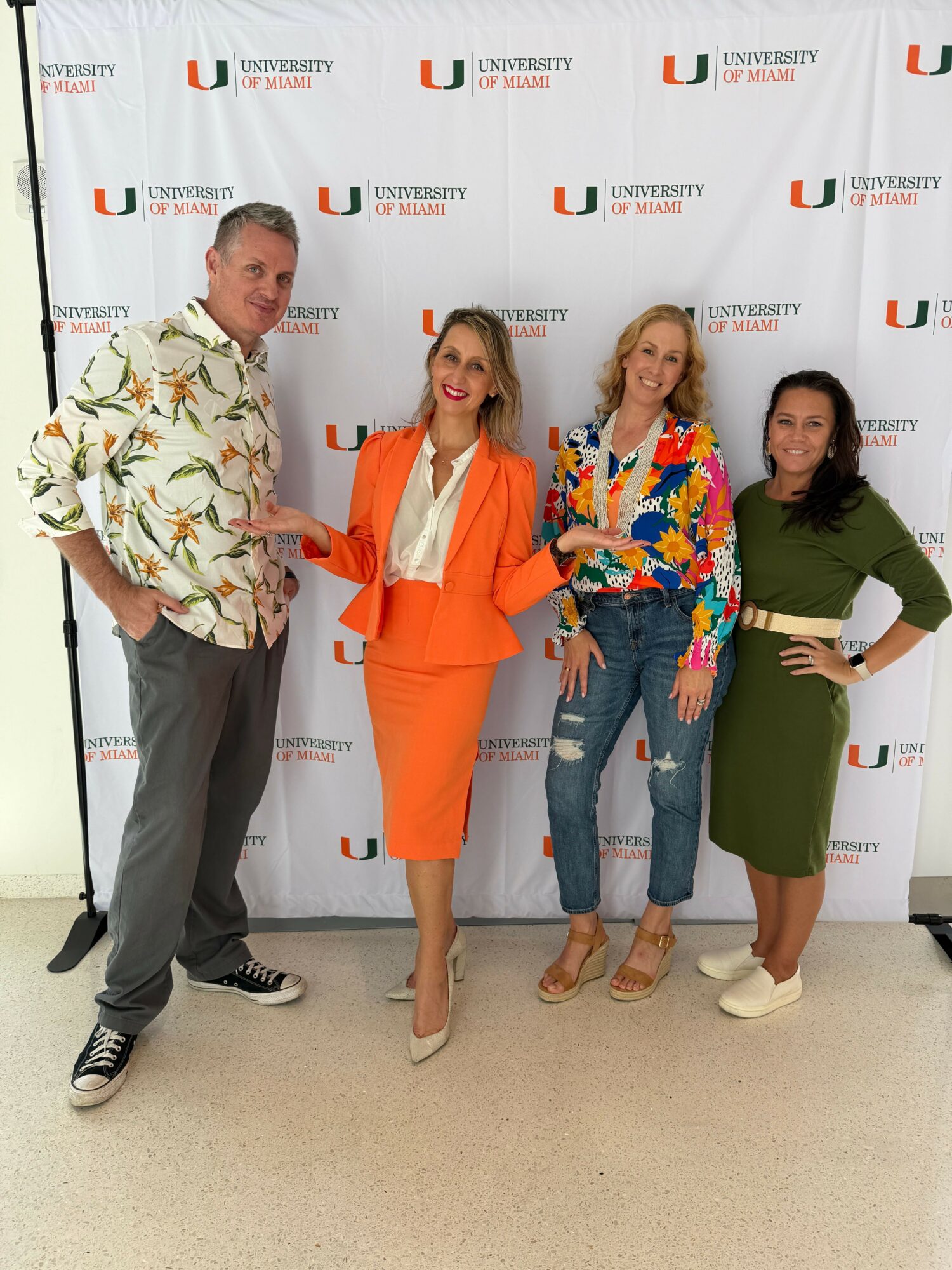 Four people standing in front of a backdrop with a logo, posing for a photo. One woman in orange dress, others in casual attire.