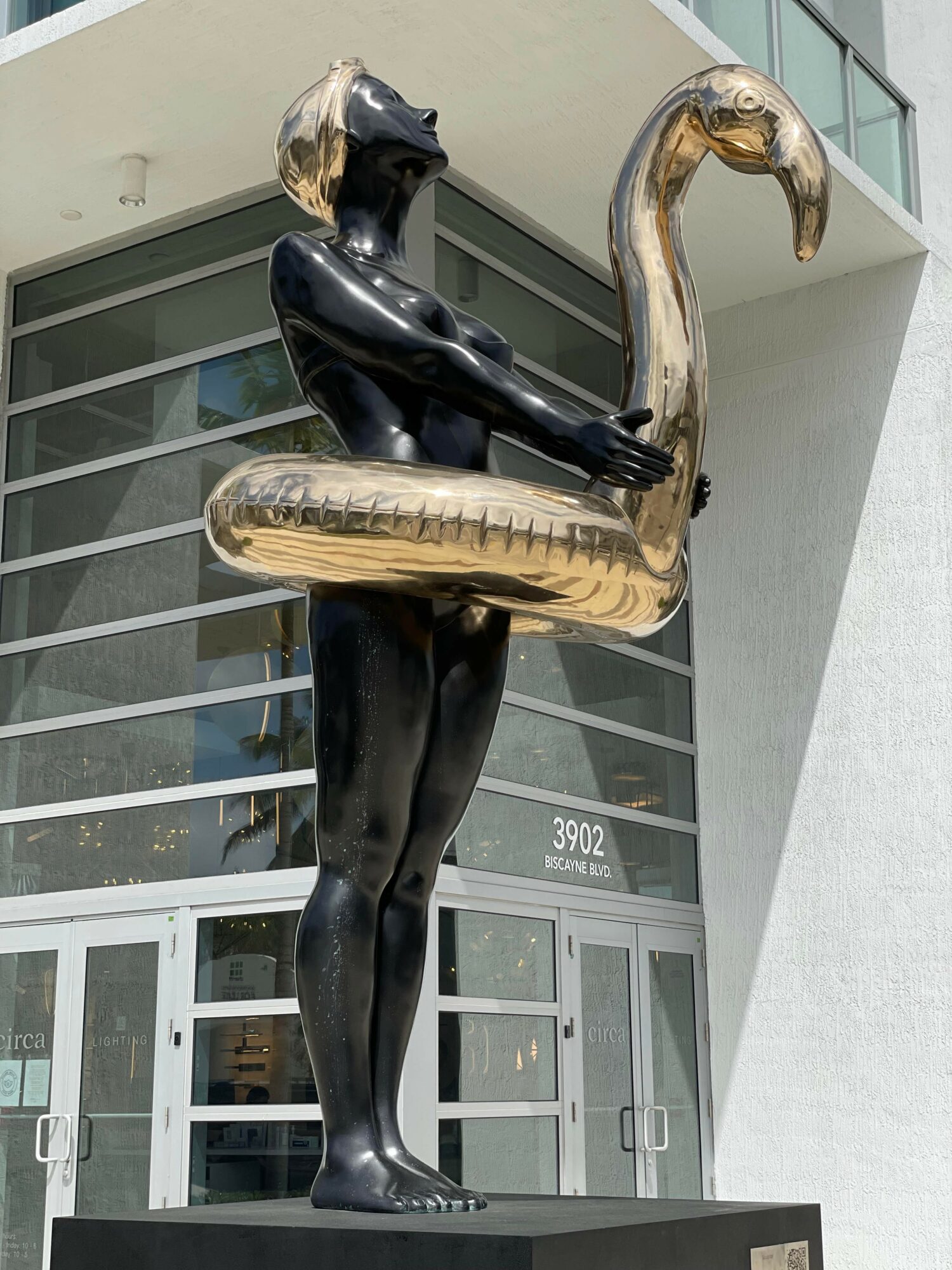 Black sculpture of a woman holding a large inflatable swan, standing outside a modern building with glass windows.