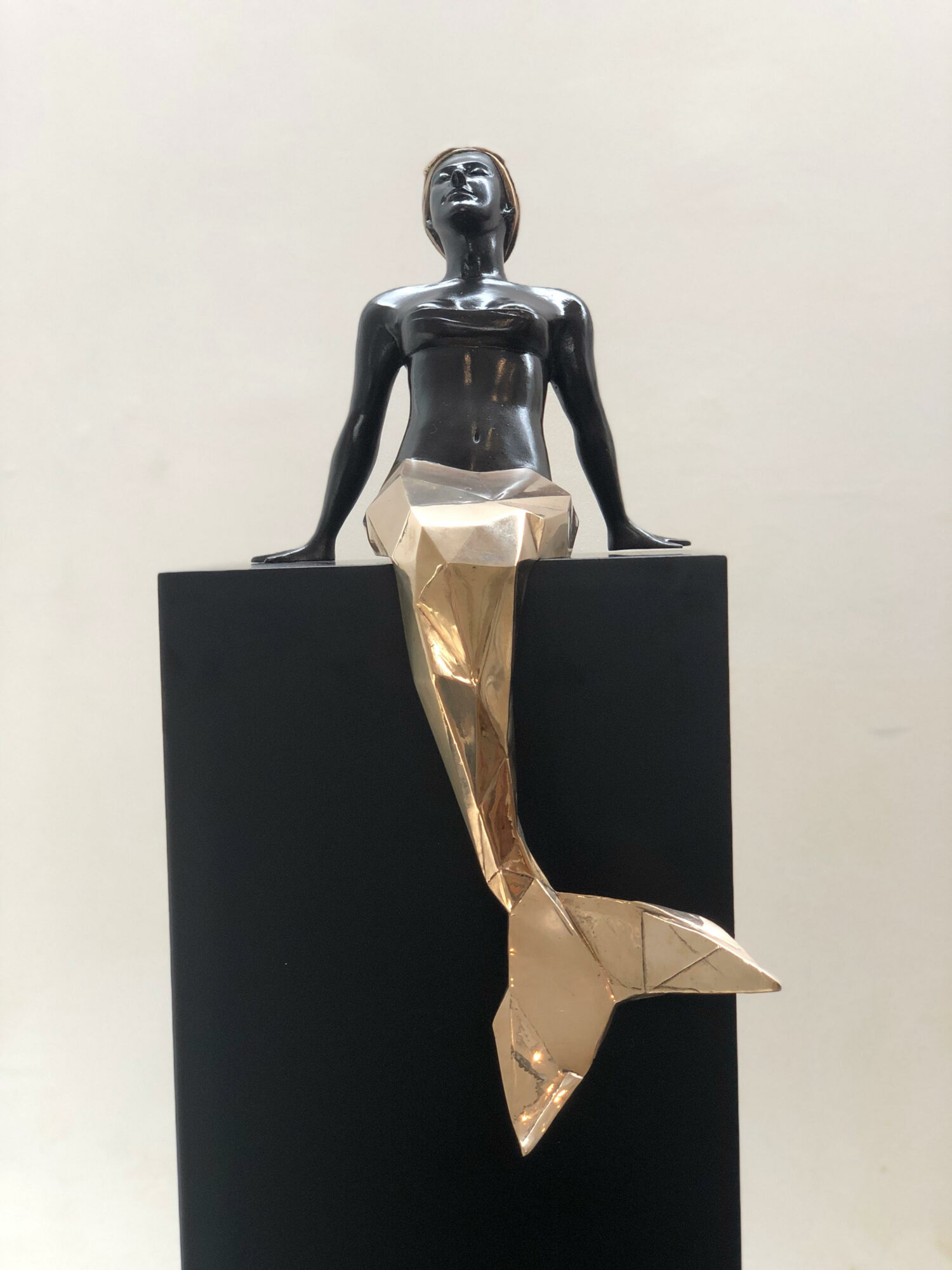 Sculpture of a woman with a metallic finish, sitting on a black block with a gold-colored tail extending downward.