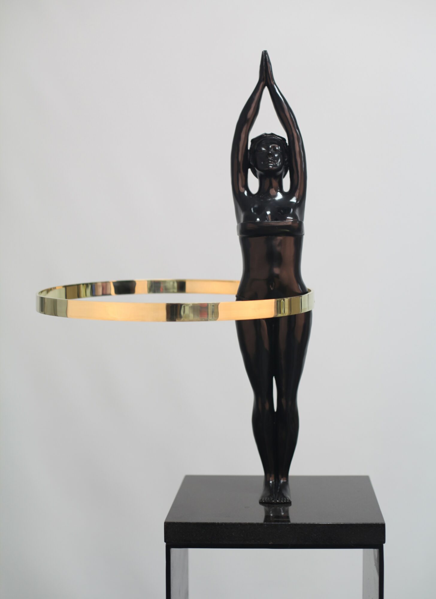 Black sculpture of a person with arms raised above head, standing on a platform with a circular ring around waist.