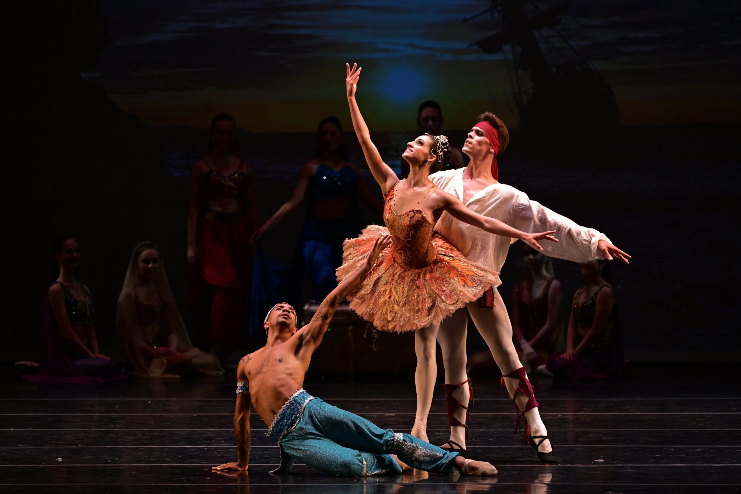 Three ballet dancers perform on stage with others in the background, one dancer in a tutu is lifted by two others.