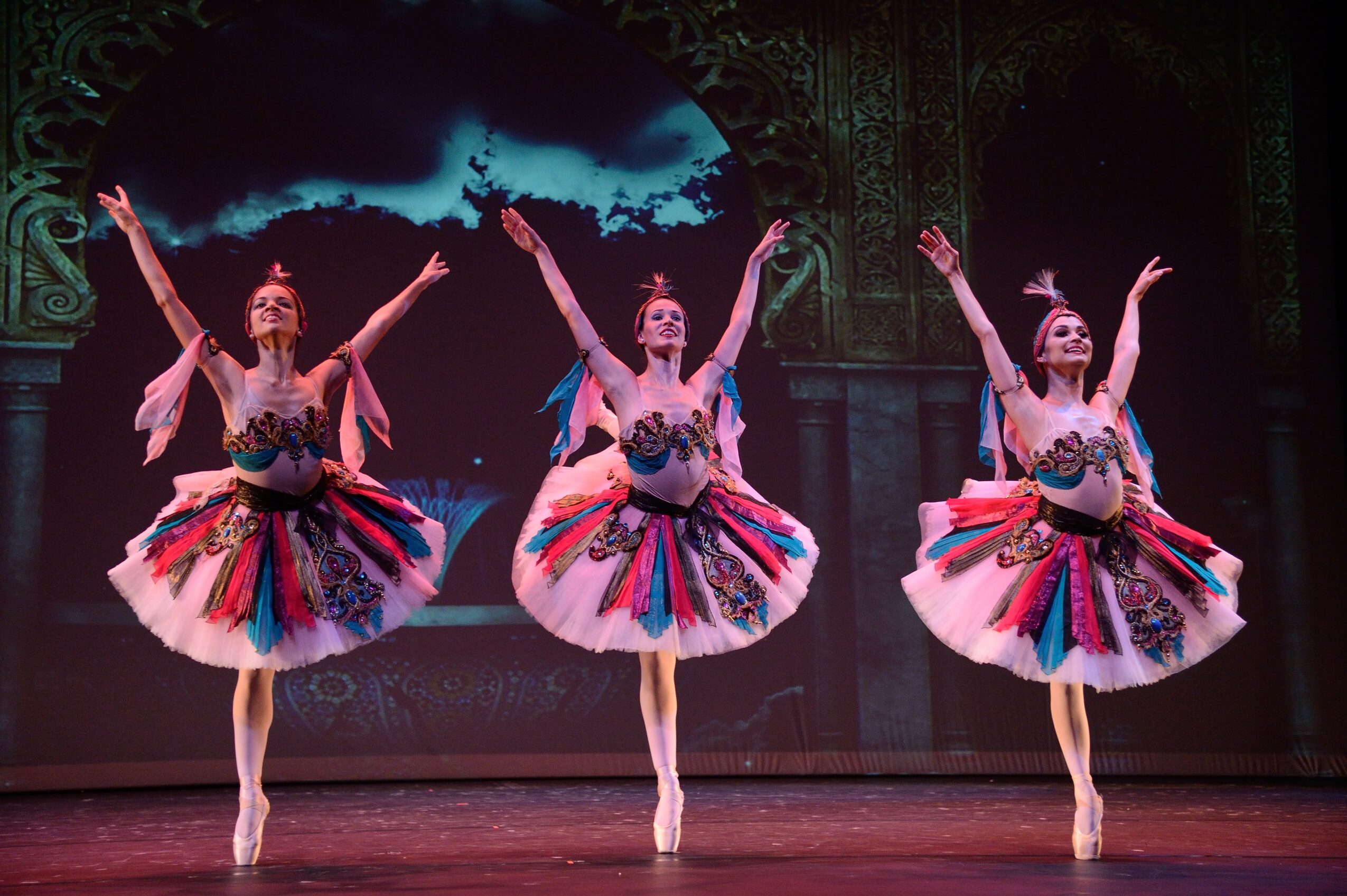 Three ballet dancers in colorful tutus perform on stage with arms raised, background scenery visible behind them.