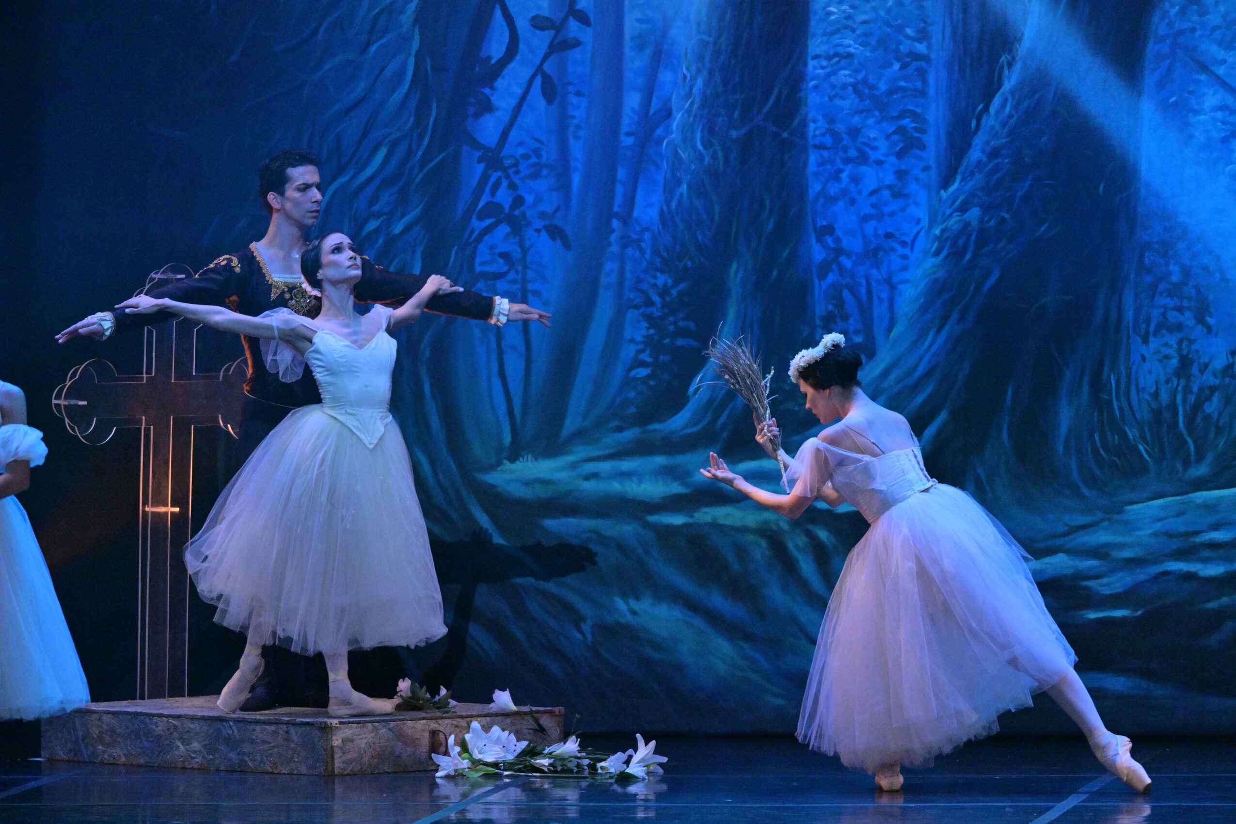 Three ballet dancers perform on stage with a forest backdrop, two standing and one kneeling, all in white tutus.
