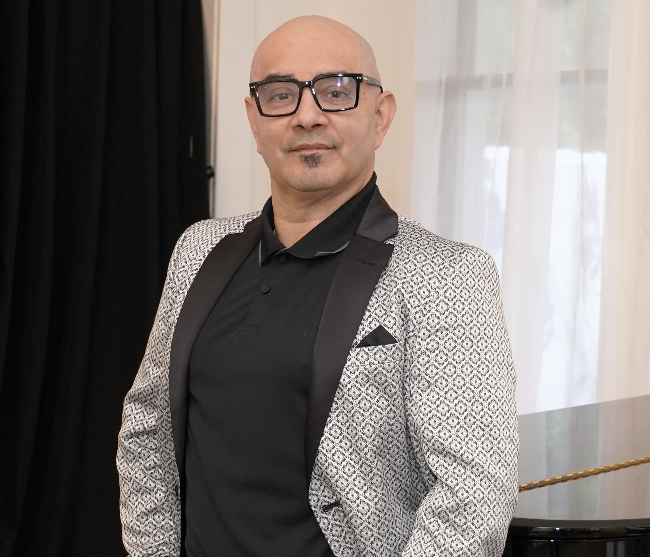 Bald man with glasses in a patterned blazer and black shirt, standing indoors near a window with curtains.