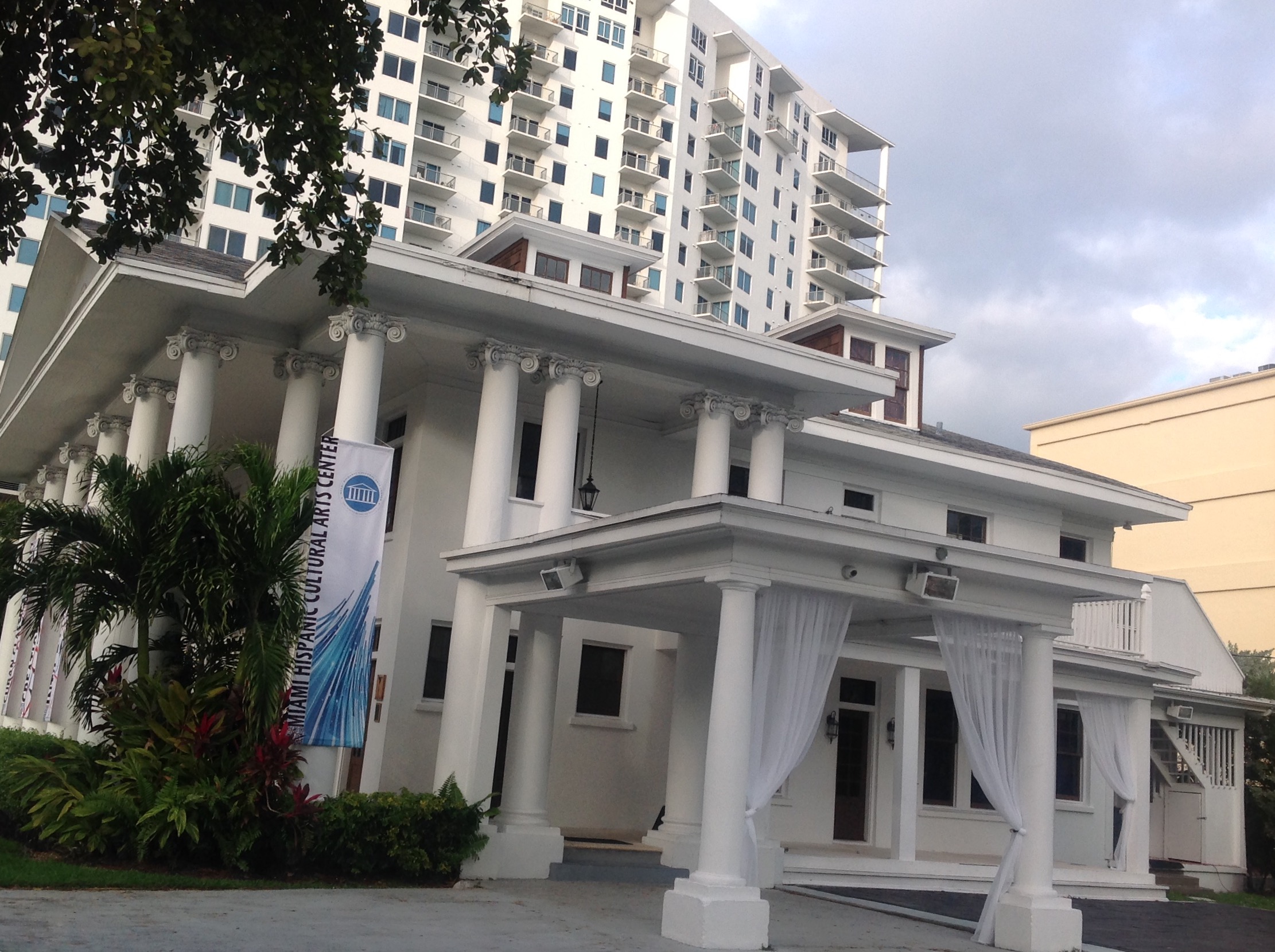 White building with columns and curtains, surrounded by greenery, with tall modern buildings in the background.