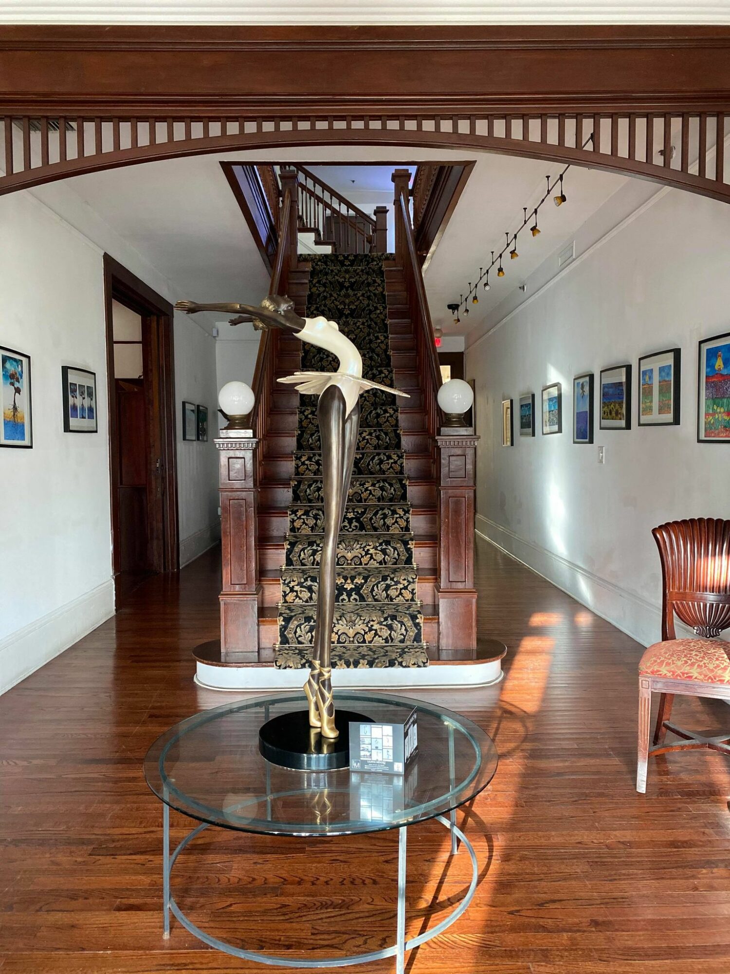 Interior view of a hallway with a staircase, framed pictures on walls, and a glass table with a decorative bird sculpture.