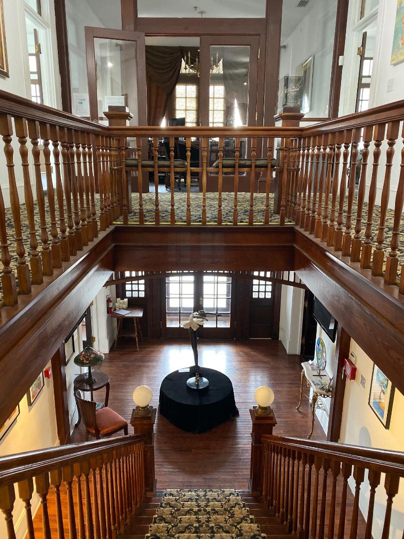 Interior view of a two-story room with a balcony, wooden staircase, and a table with a lamp in the center.