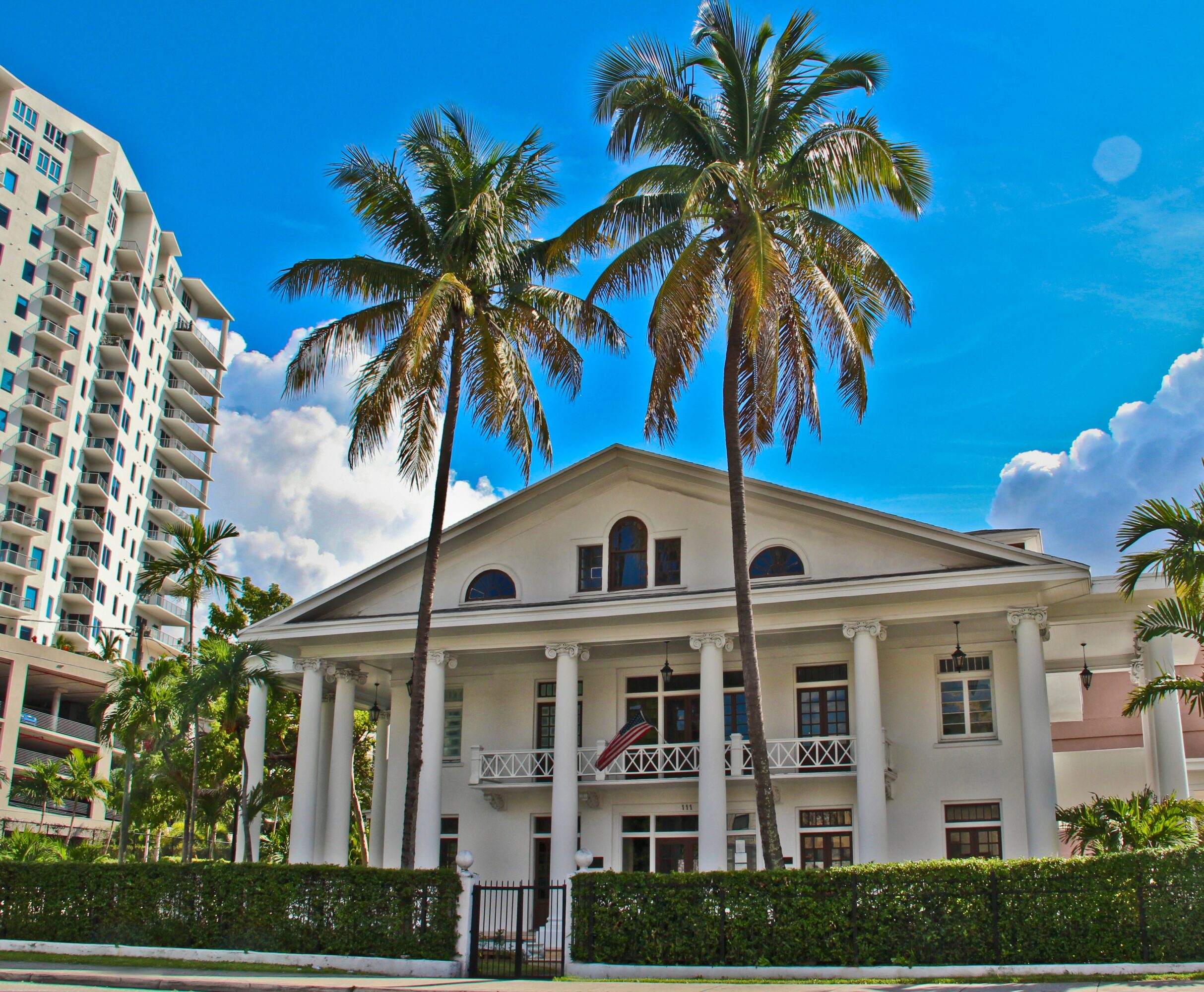 White colonial-style house with columns, surrounded by palm trees, under a blue sky with clouds.