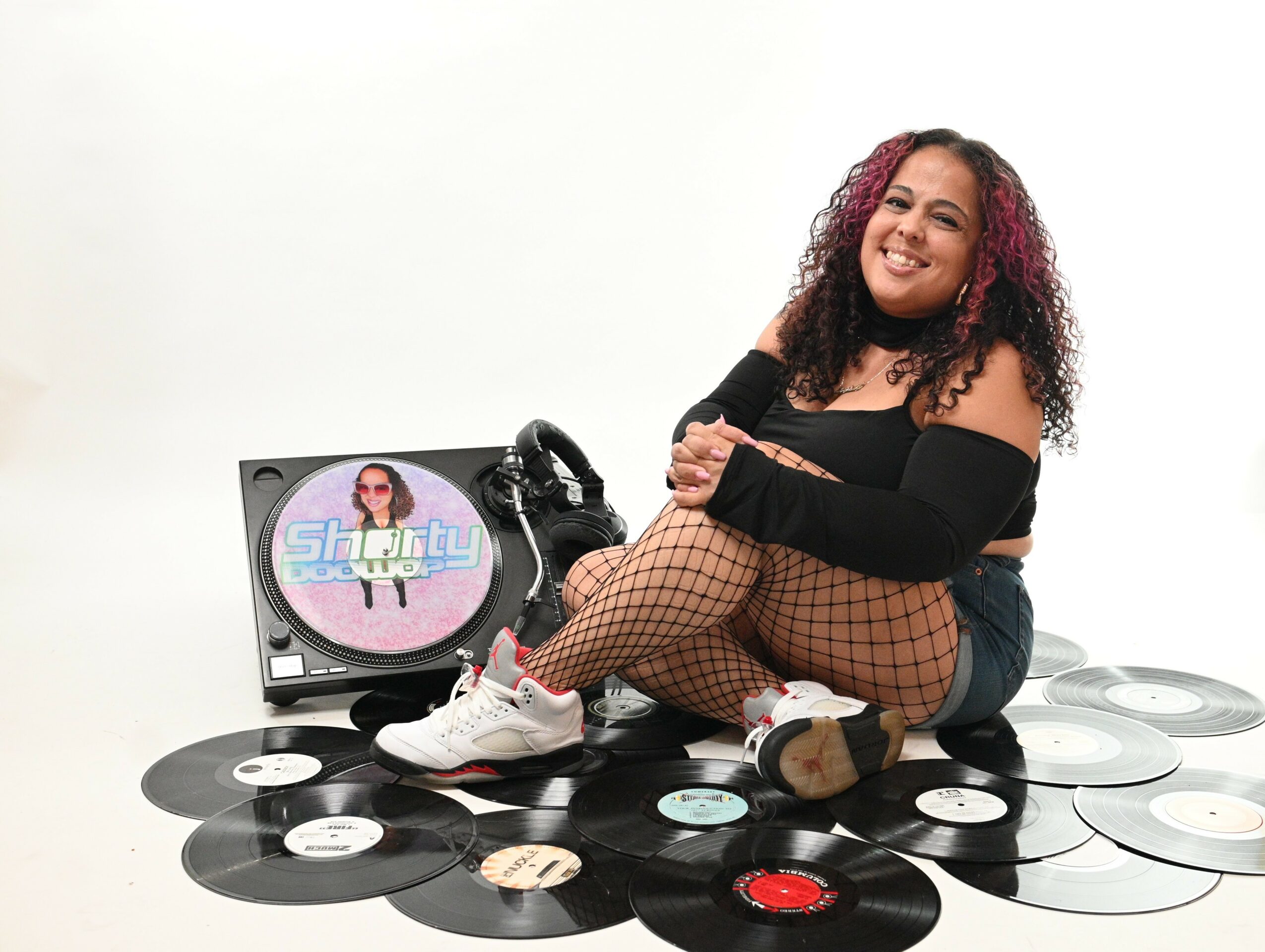 Person sitting on vinyl records with a turntable displaying a colorful logo, smiling, wearing fishnet stockings and casual clothes.