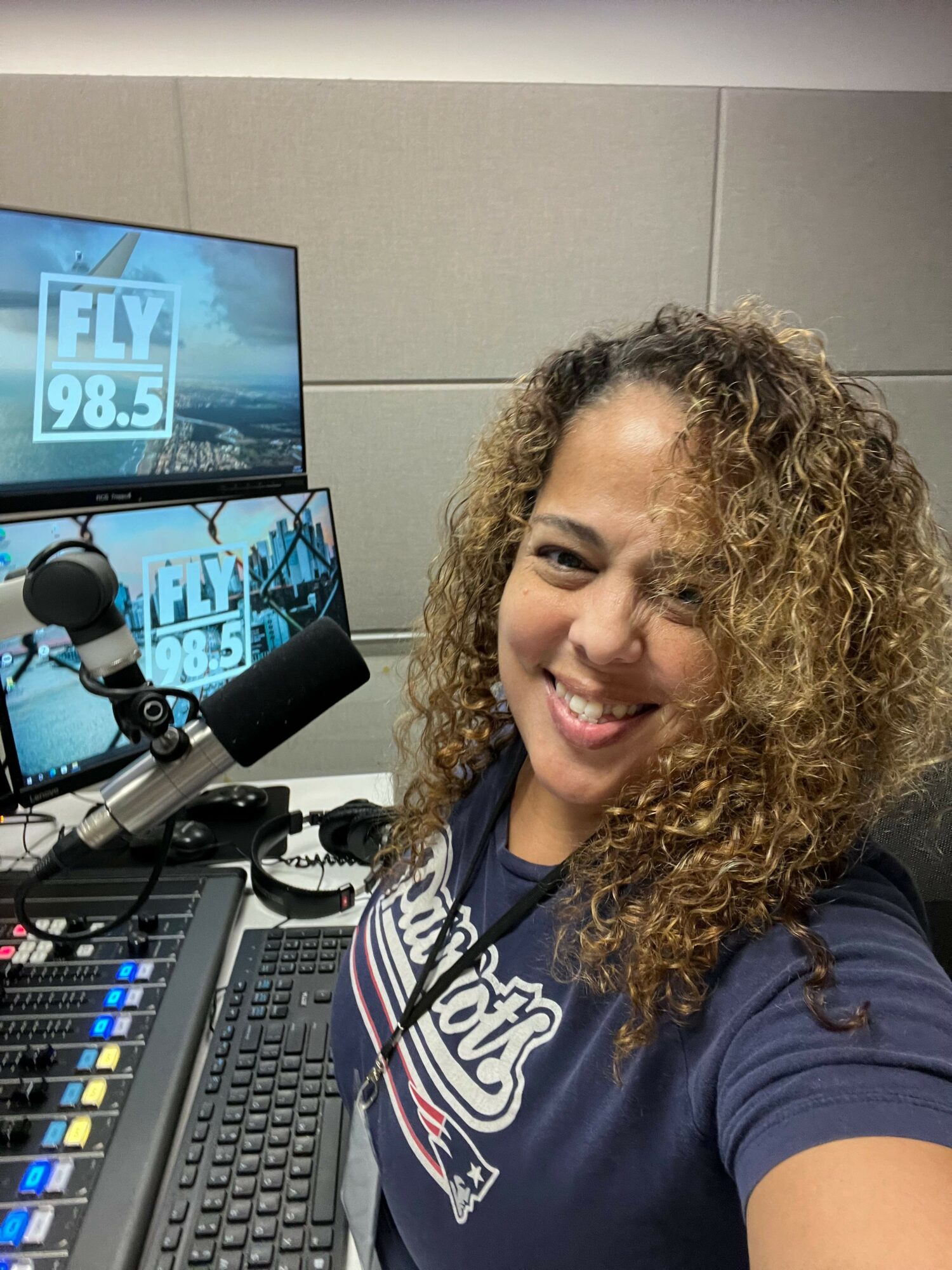 Woman with curly hair smiling in a radio studio, with audio equipment and a computer monitor visible.