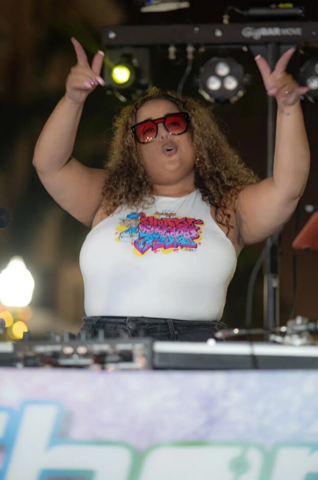 Woman with curly hair wearing sunglasses and a colorful graphic tank top, raising hands at a DJ booth outdoors.
