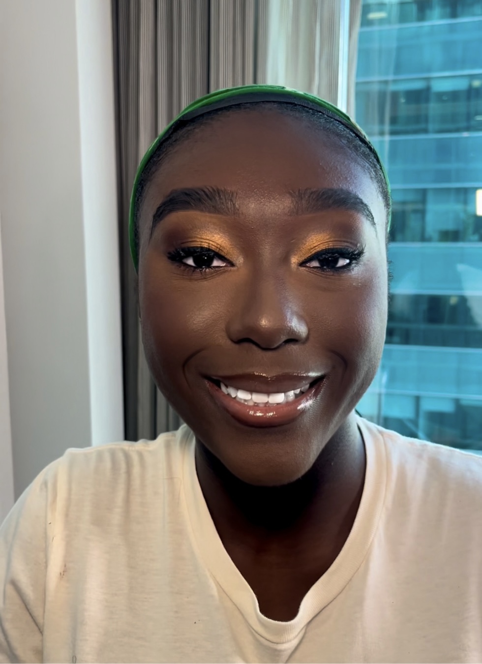 Smiling woman with dark skin, wearing a green headband and white shirt, indoors near a window with city view.