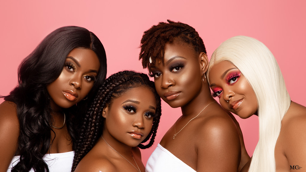 Four women with different hairstyles and makeup pose against a pink background.