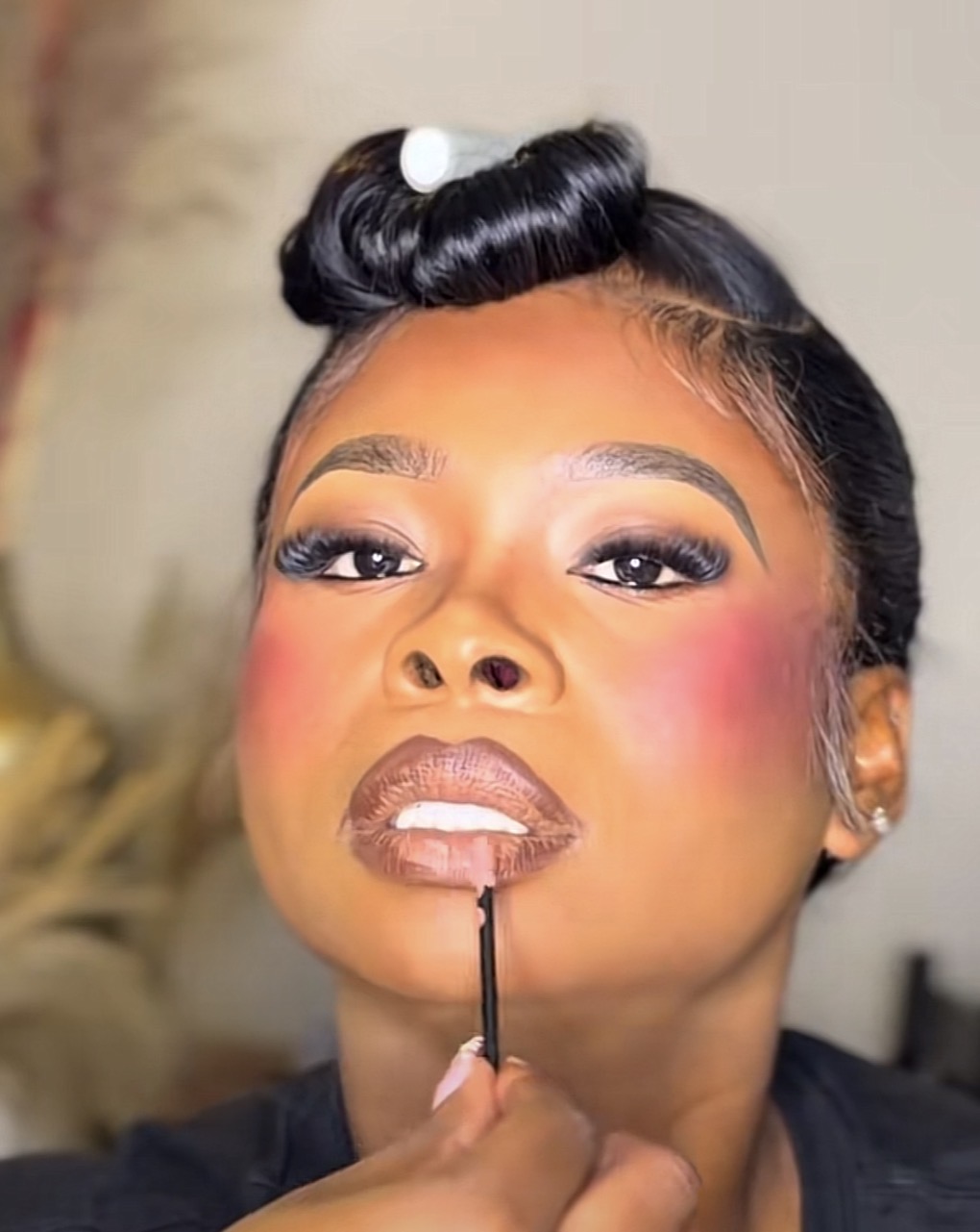 Close-up of a woman with styled hair, makeup, and a makeup brush near her lips, looking at the camera.