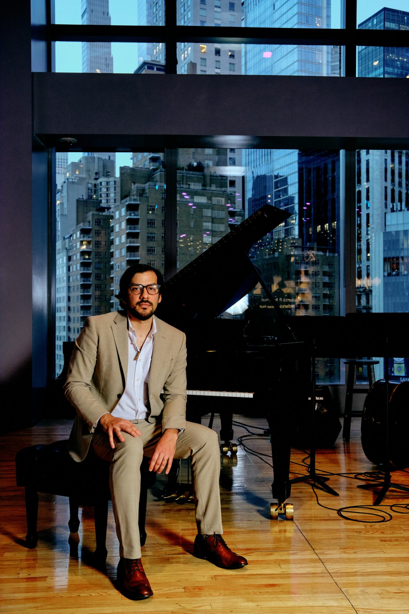 Man in beige suit sitting on a piano bench in a modern city office with large windows and a grand piano.