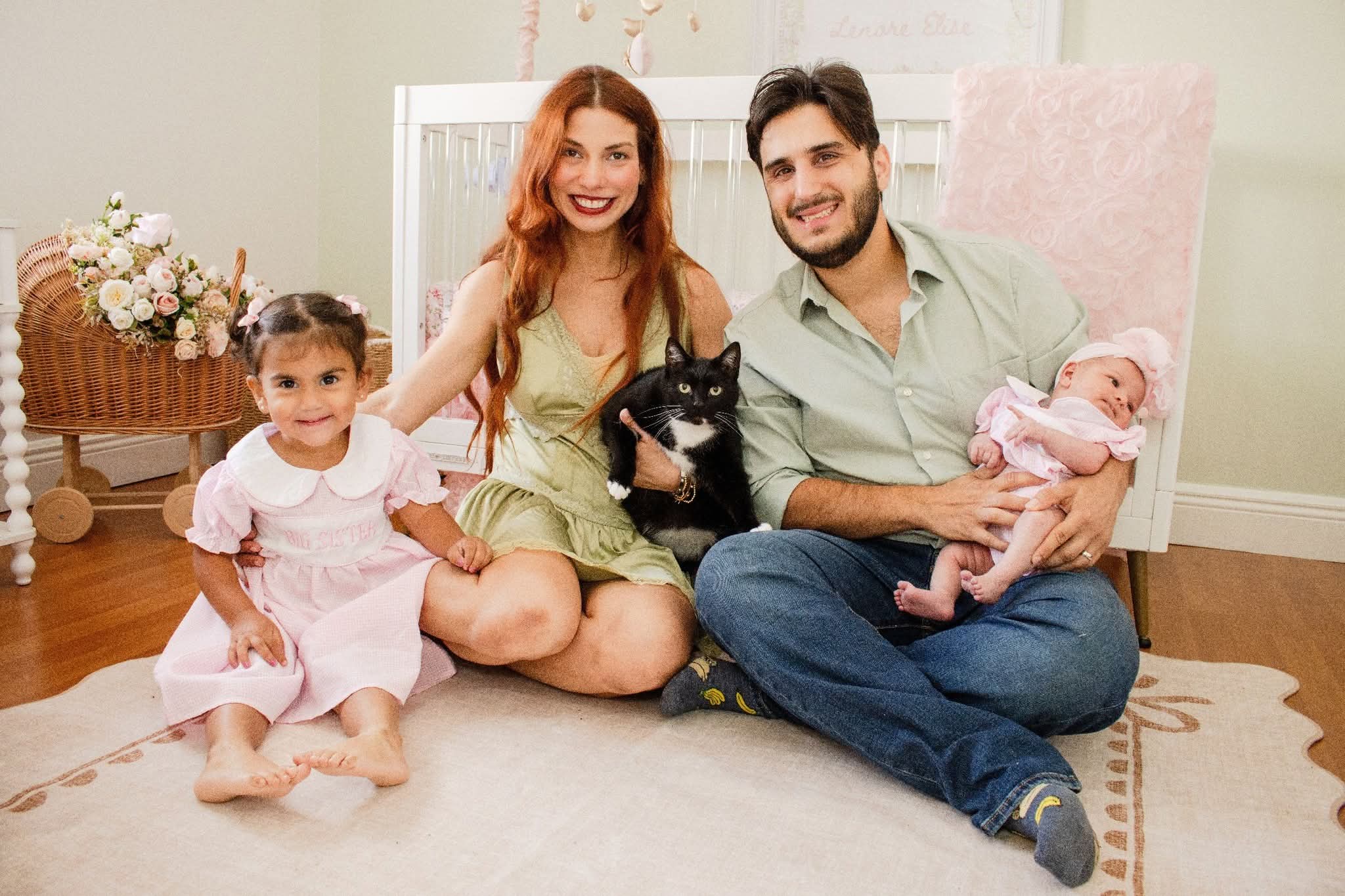 Family of four with a dog and baby sitting on the floor in a living room, smiling at the camera.