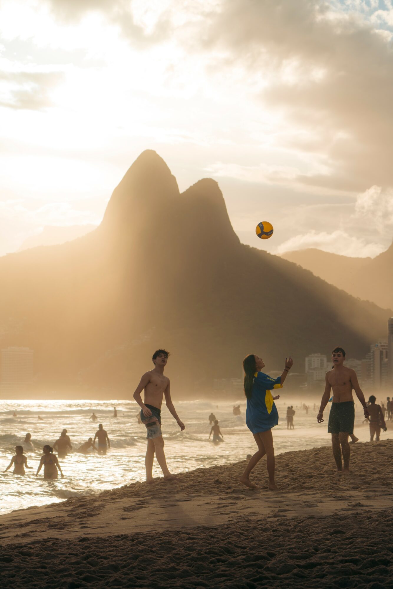 People playing volleyball and swimming on a beach with mountains and cloudy sky in the background.