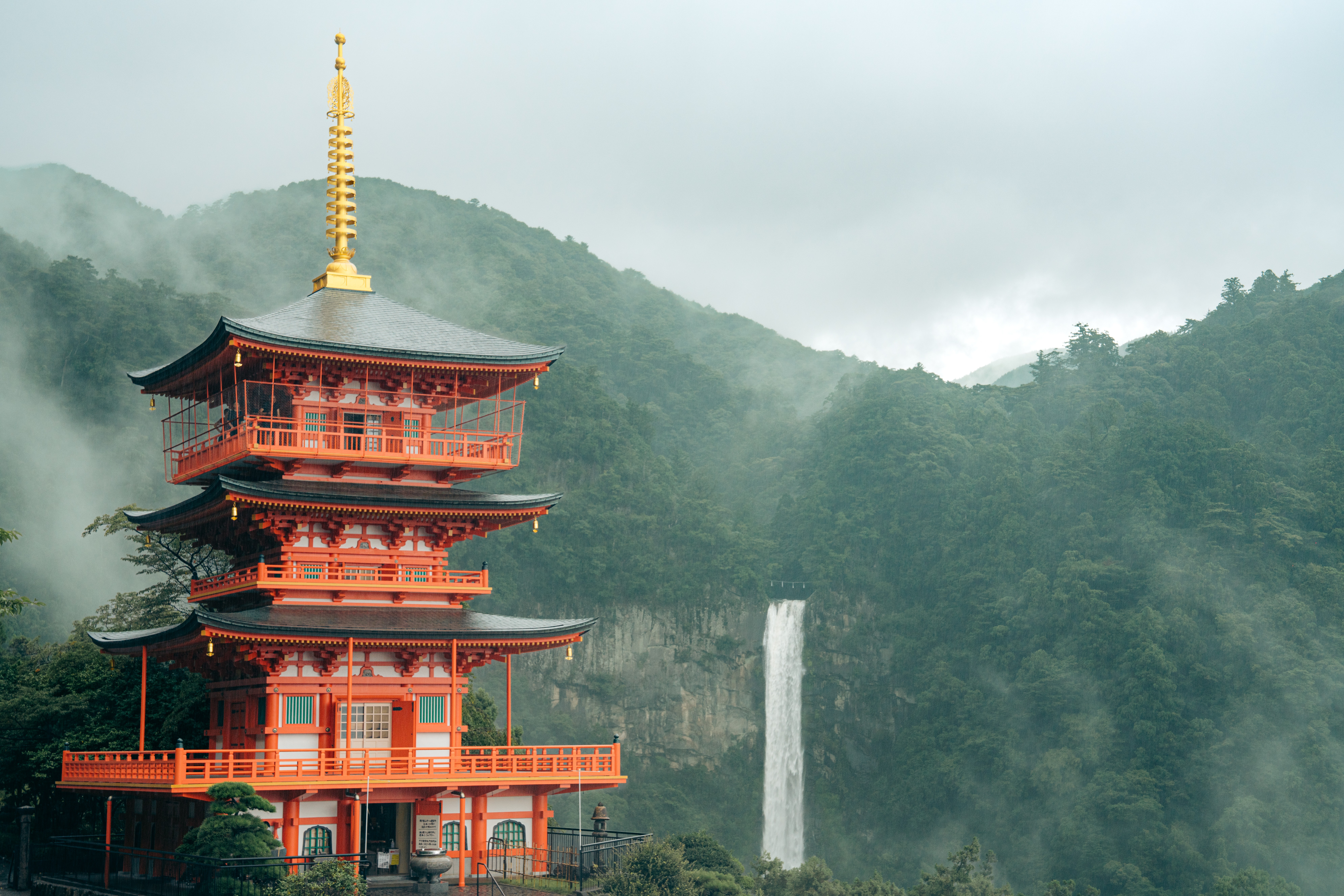 A traditional multi-story pagoda with red and black colors, set against a misty mountain landscape with a waterfall in the background.