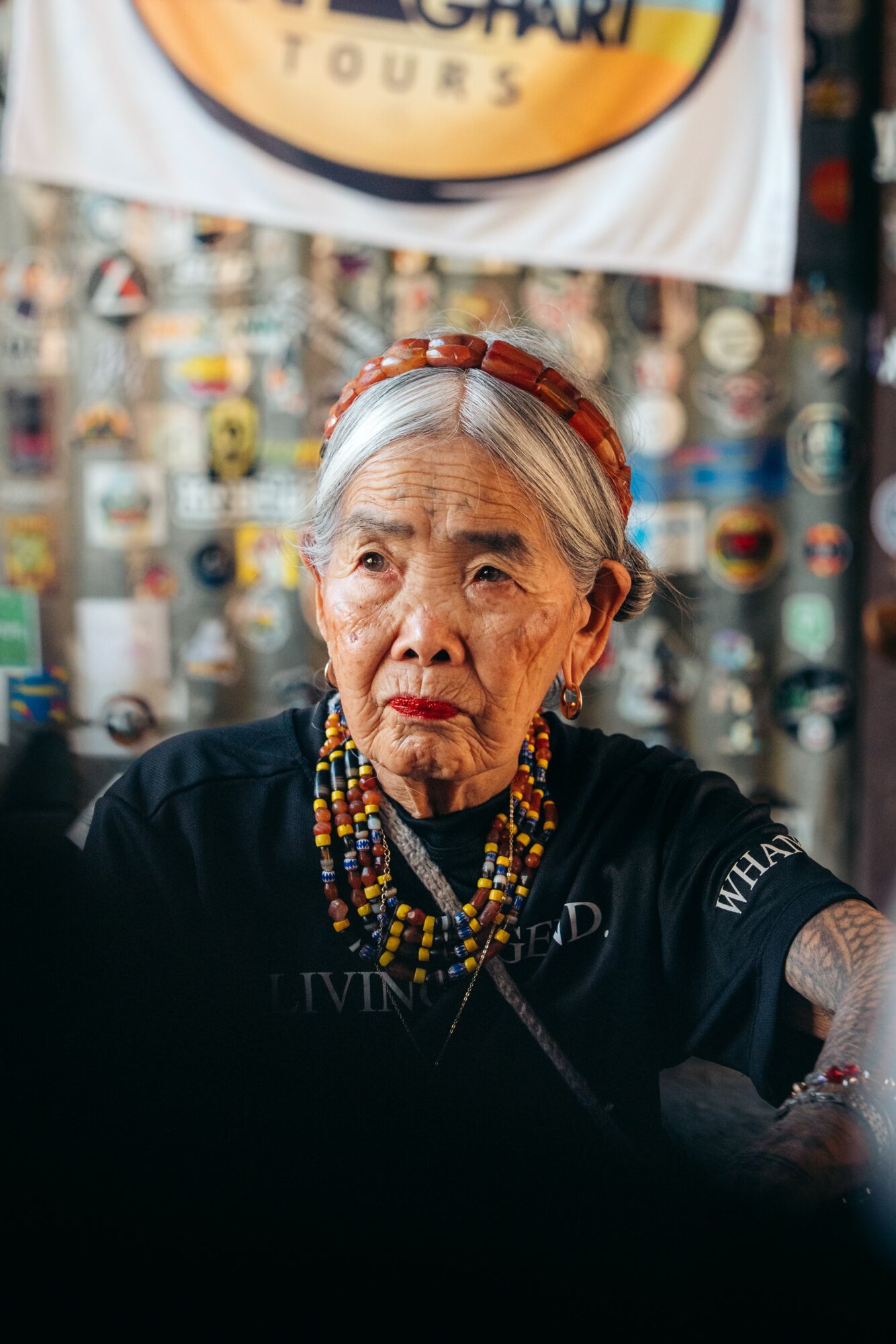 Elderly woman with gray hair wearing colorful beaded necklaces and a headband, sitting indoors with a patterned background.
