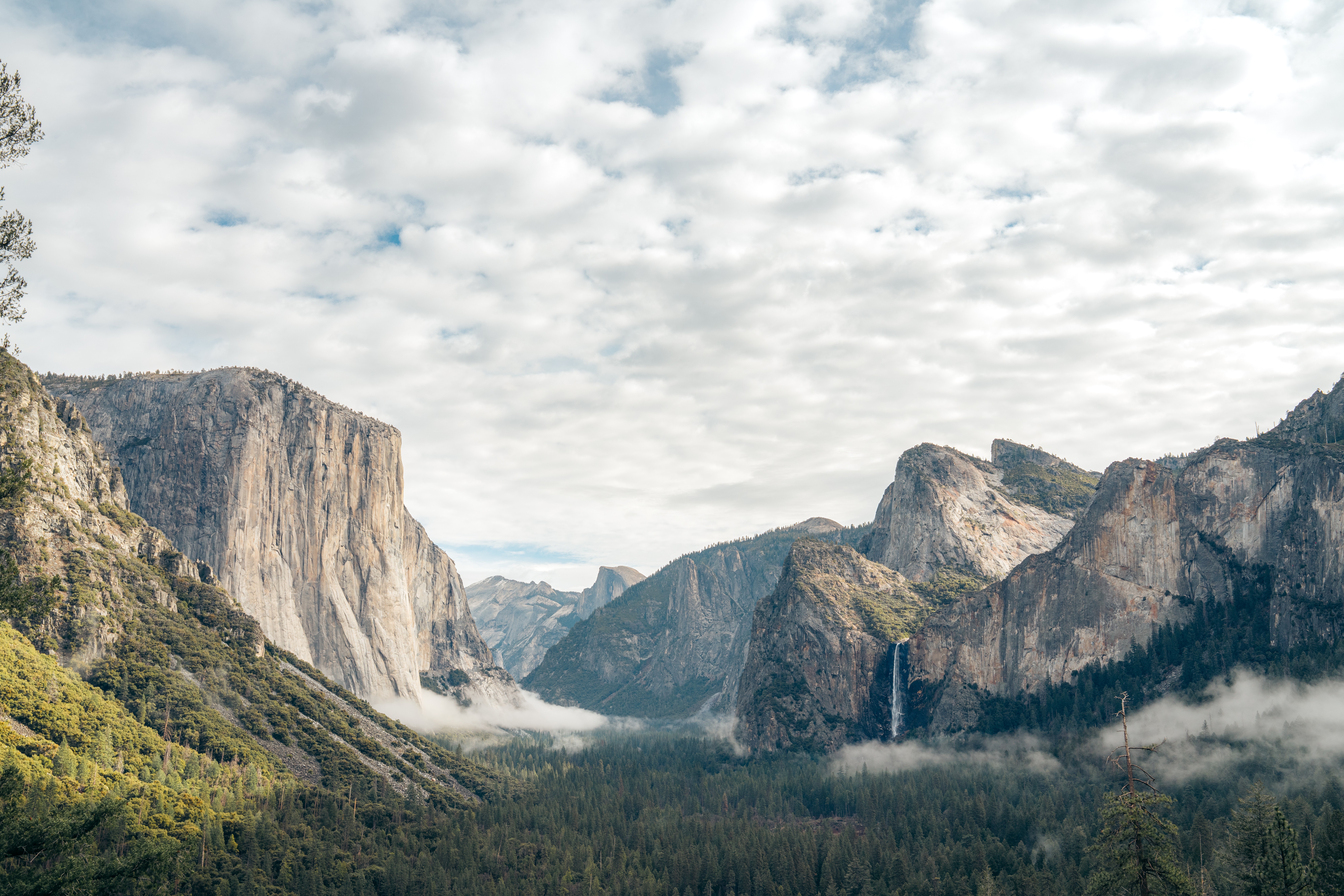 Mountain landscape with cliffs, forest, and cloudy sky, viewed from a low angle.