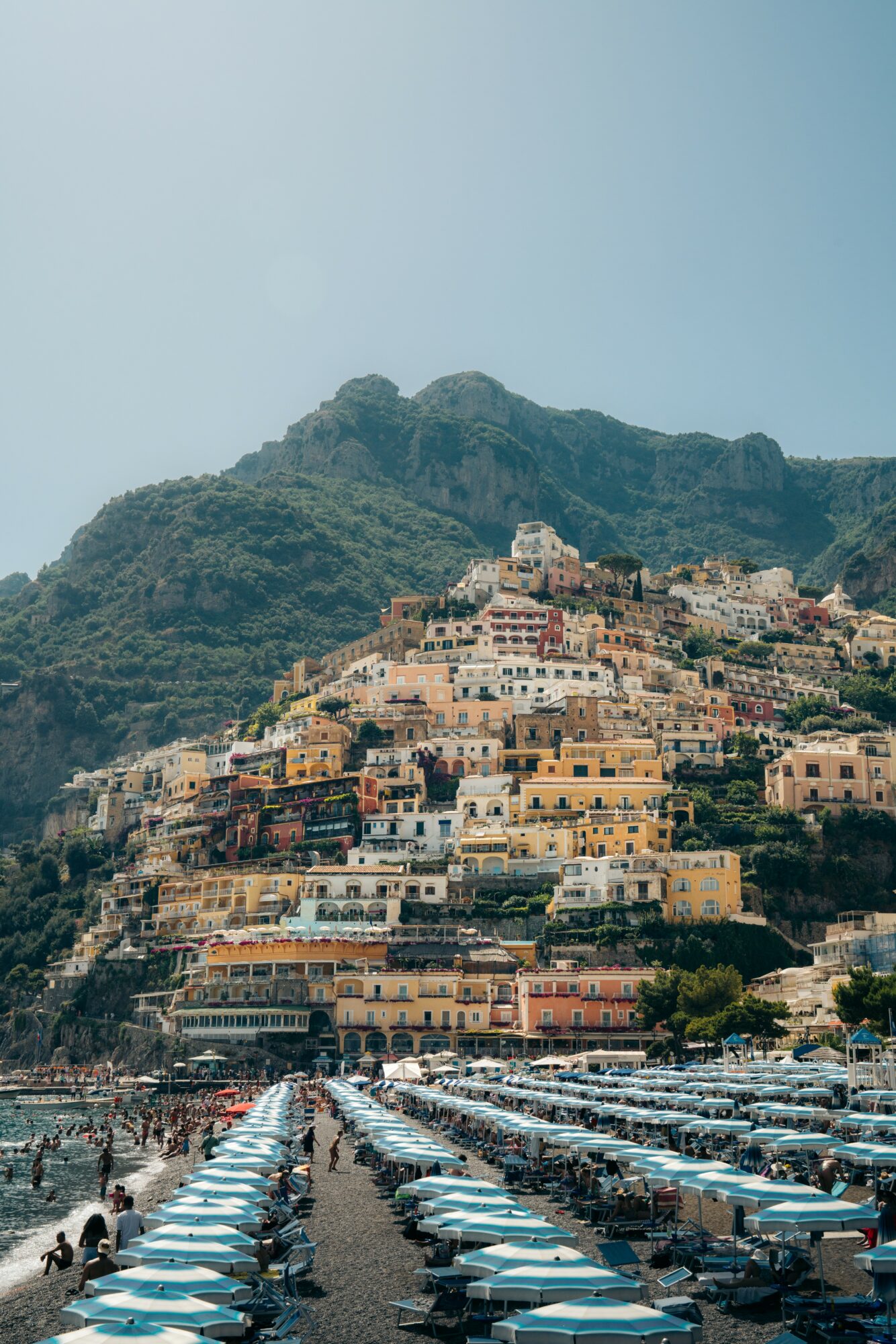 Coastal town with colorful buildings on a hillside, beach with umbrellas and people, mountain in background, clear sky.