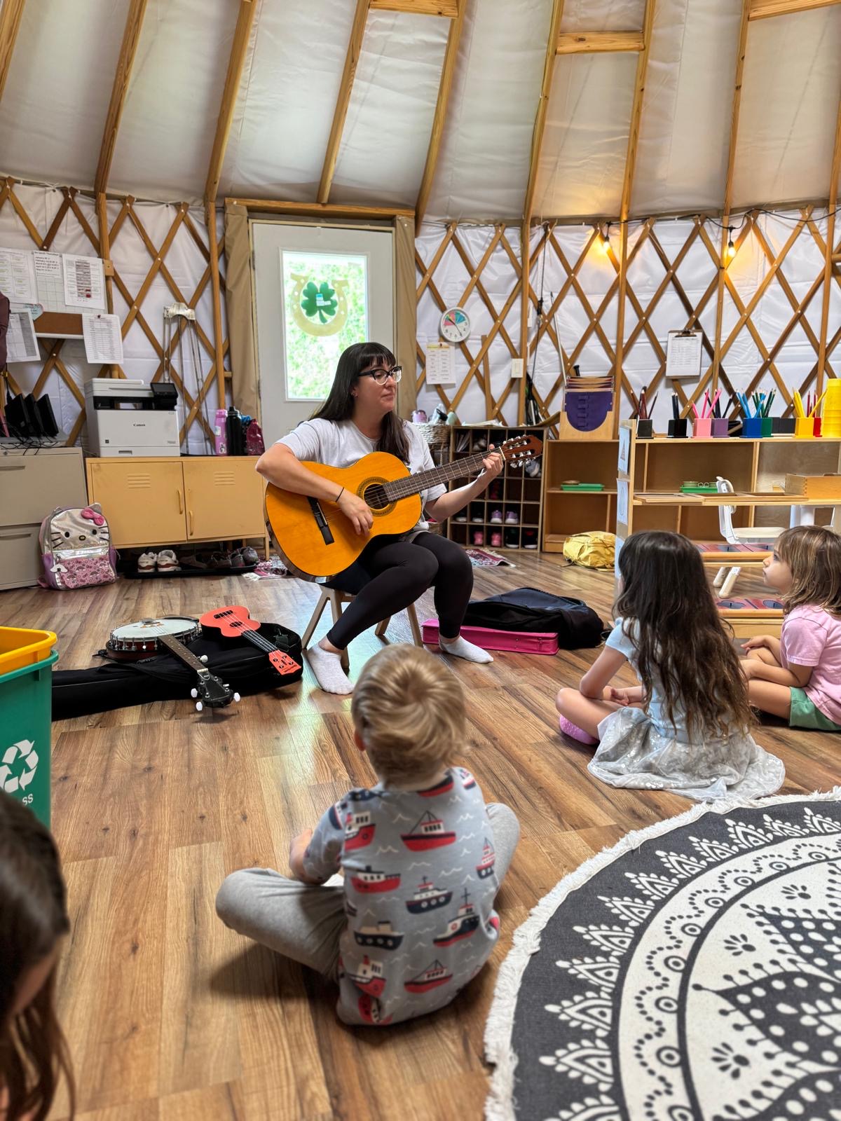 Girl playing guitar for children in a yurt-like room with wooden walls and shelves, children sitting on the floor listening.