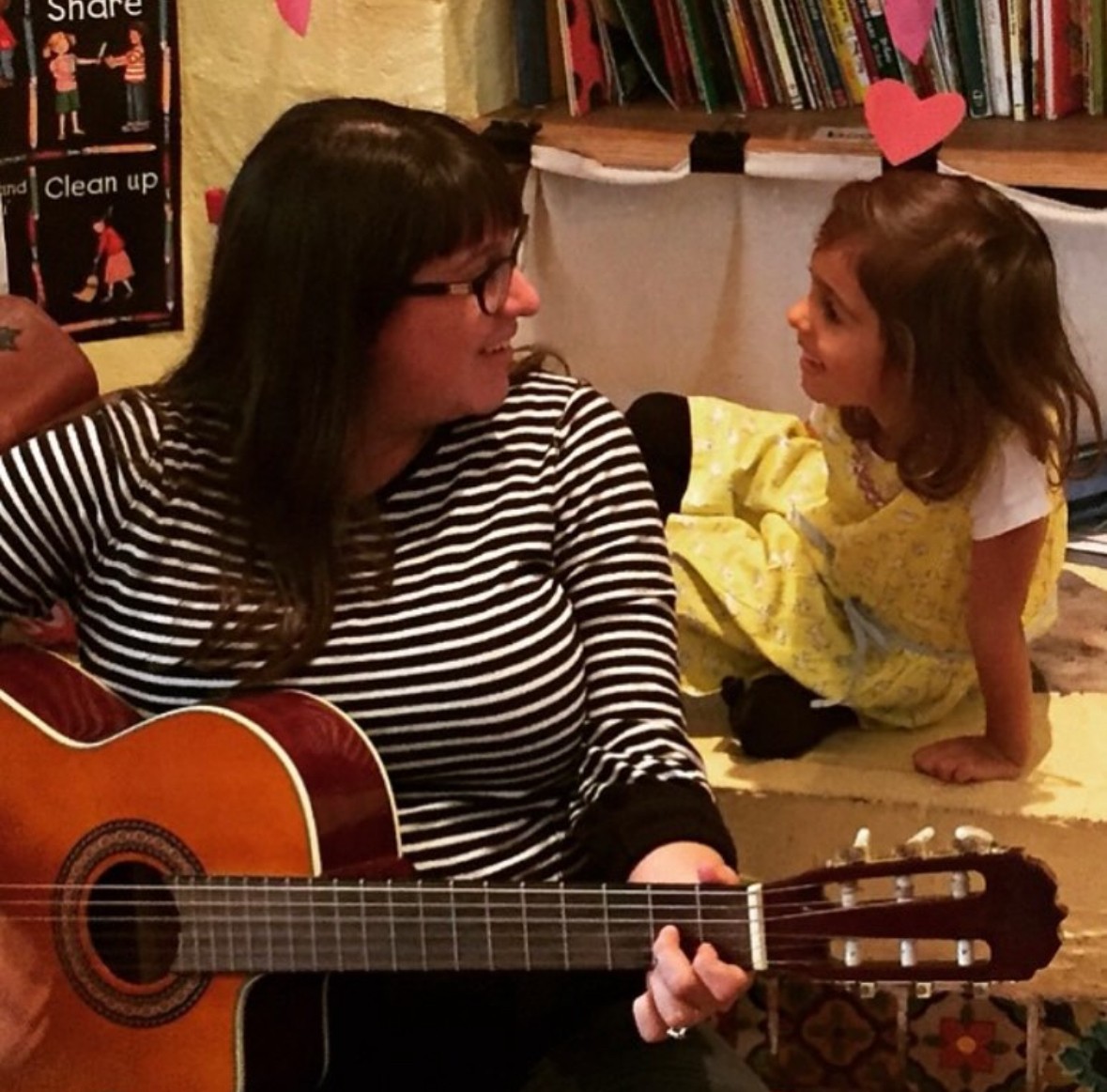 Woman playing guitar and a girl looking at her, with books and a heart decoration in the background.
