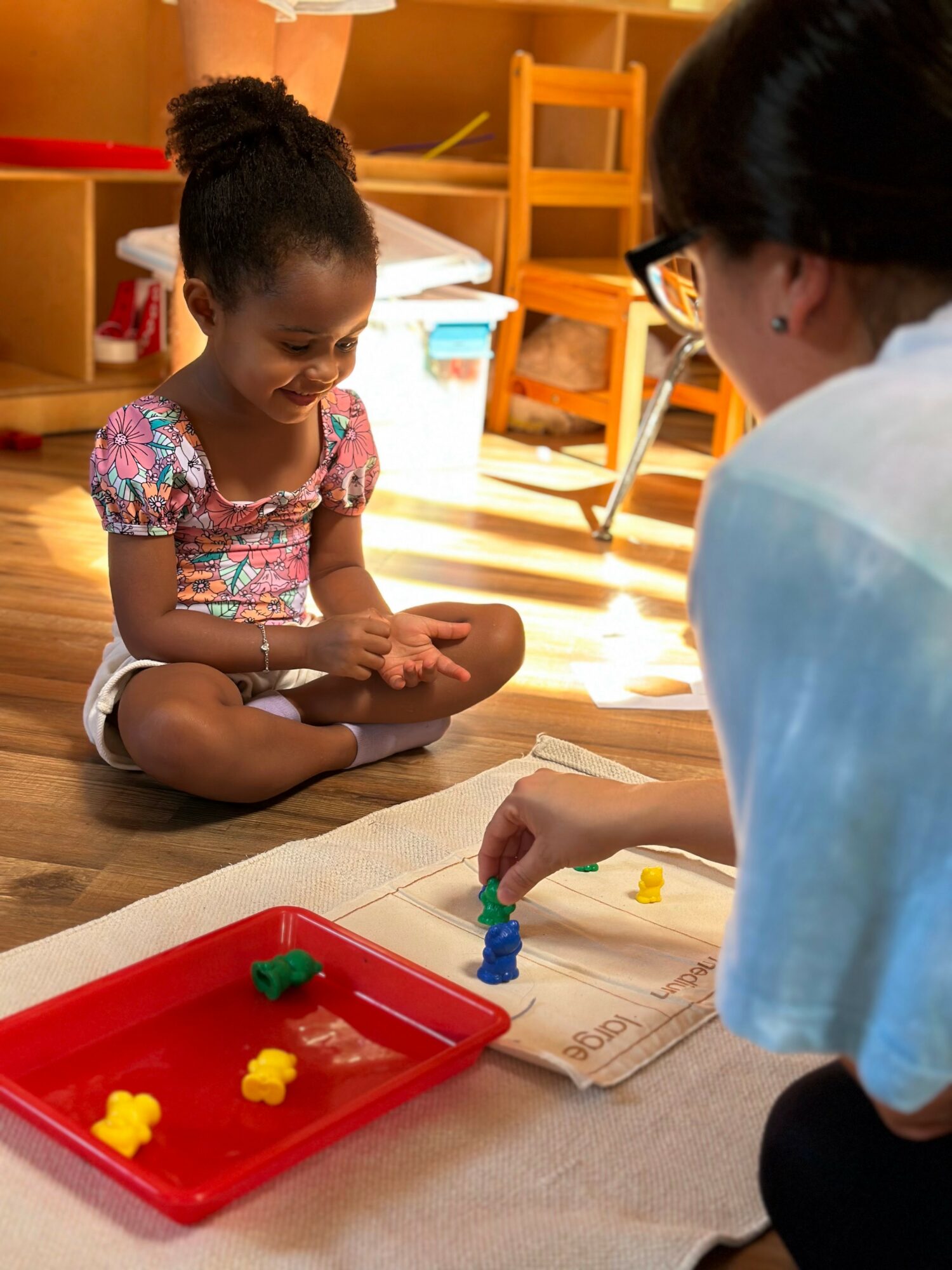 Young girl sitting cross-legged on the floor, smiling, with a woman playing a game with colorful game pieces on paper.
