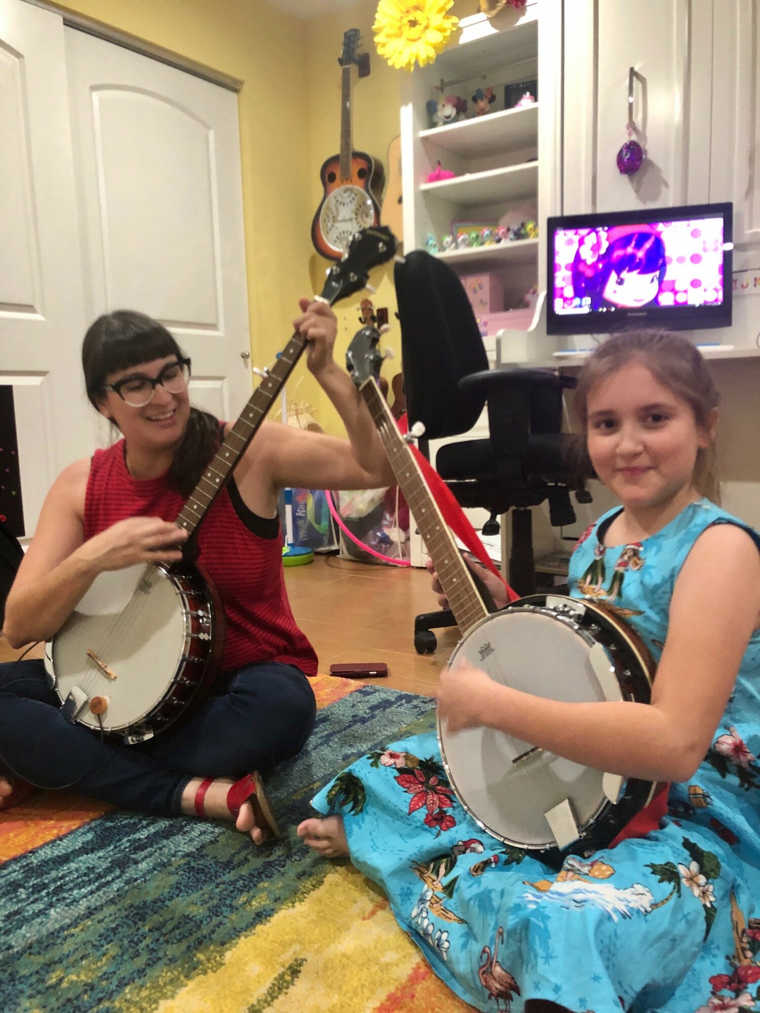 Two children sitting on a colorful rug, playing banjos in a room with a computer, shelves, and musical instruments.