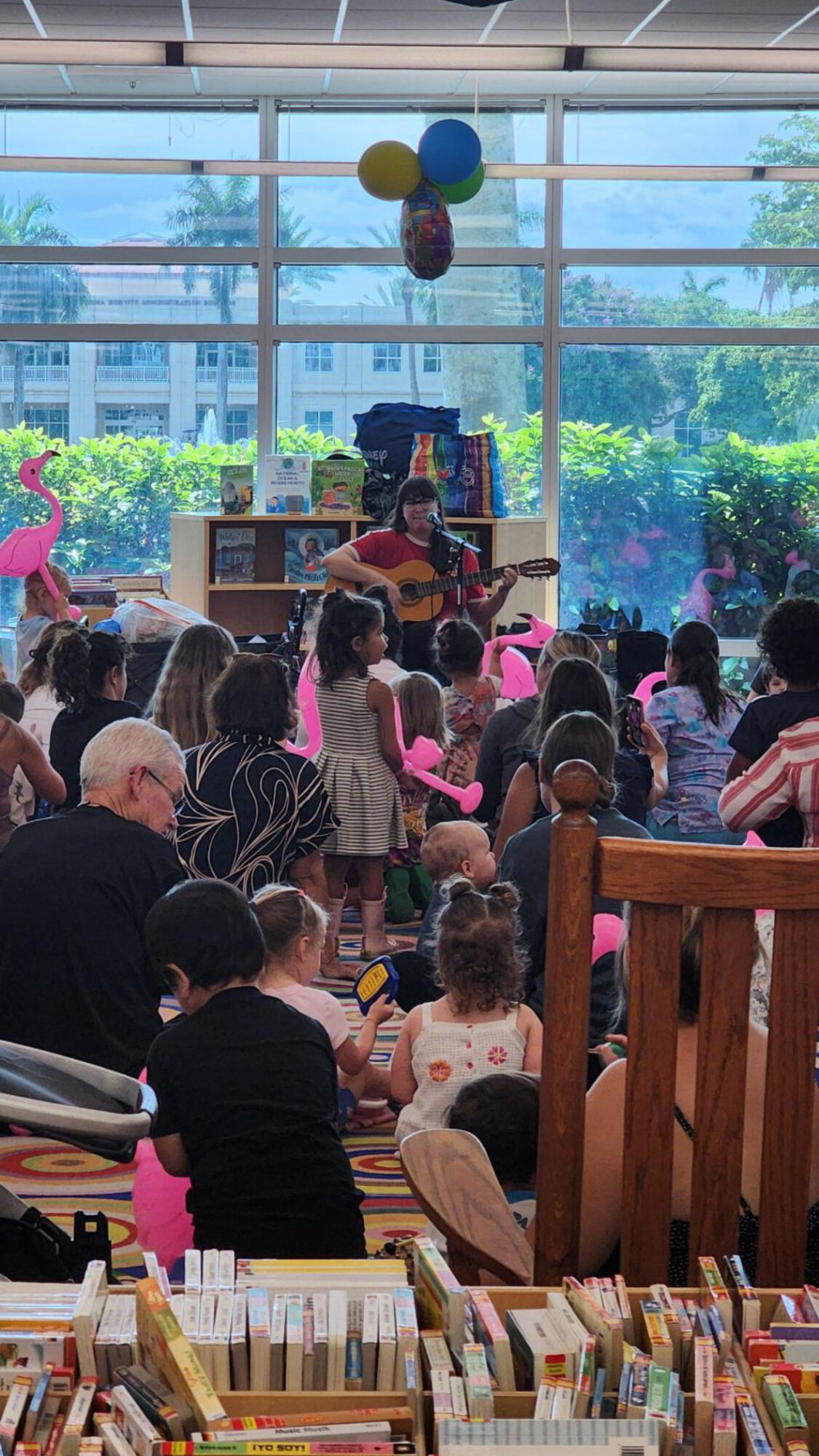 Person playing guitar on stage with balloons overhead, audience seated indoors with books in foreground.
