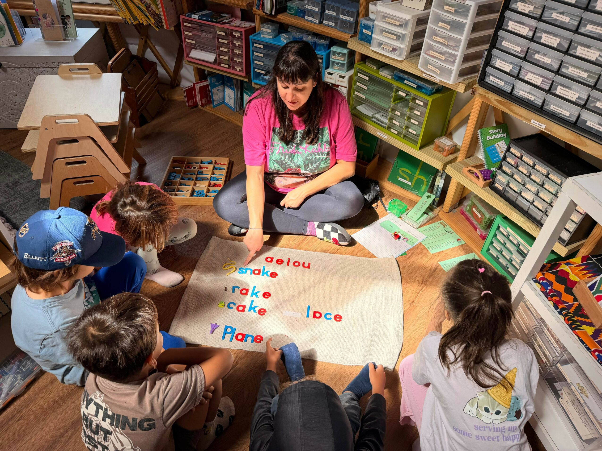 Teacher sitting on the floor with children around her, teaching words on a large paper in a classroom.