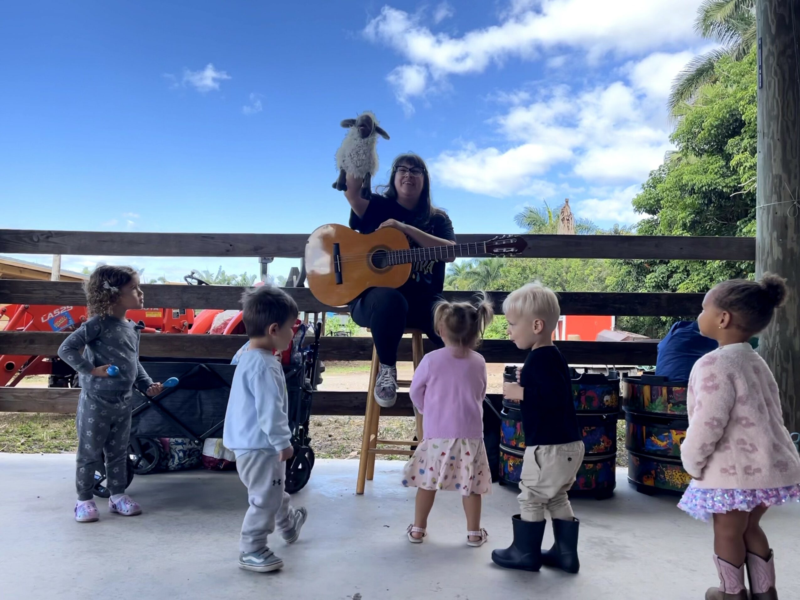 Person playing guitar with a puppet on their head, surrounded by children outdoors on a sunny day.