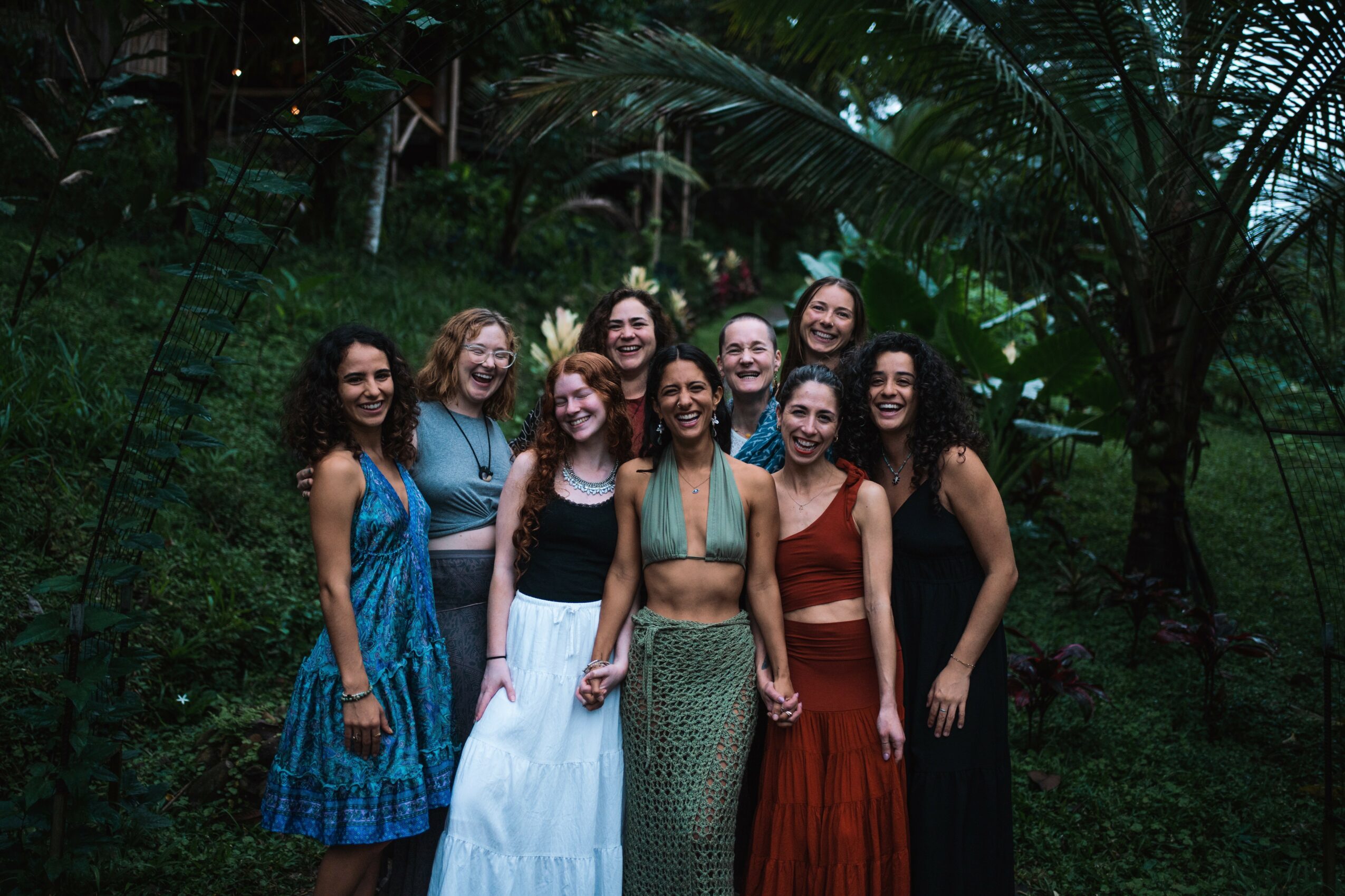 Group of nine women standing outdoors among tropical plants, smiling and posing for a photo.