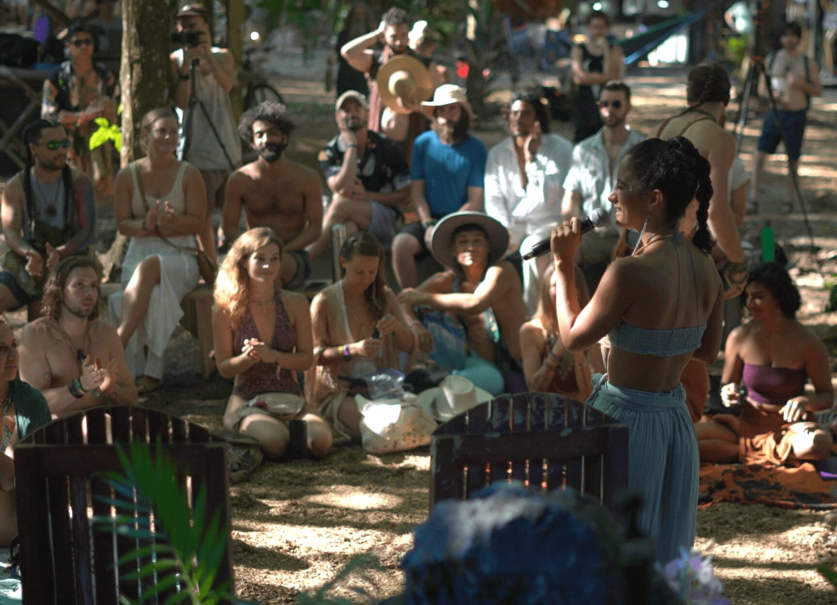 A woman performs for a seated audience outdoors, with trees and sunlight filtering through leaves.