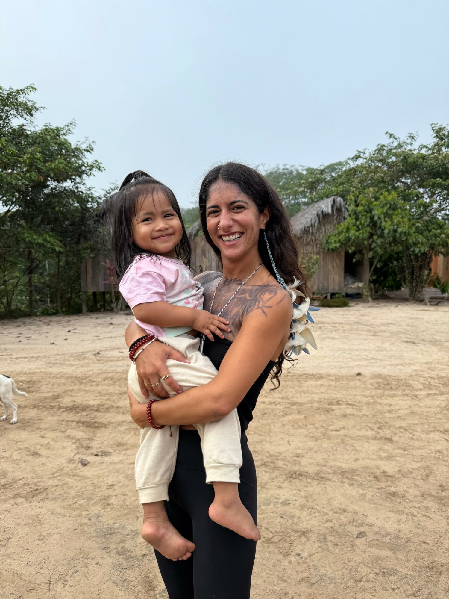 Woman holding a smiling young girl outdoors on a dirt ground with trees and a building in the background.