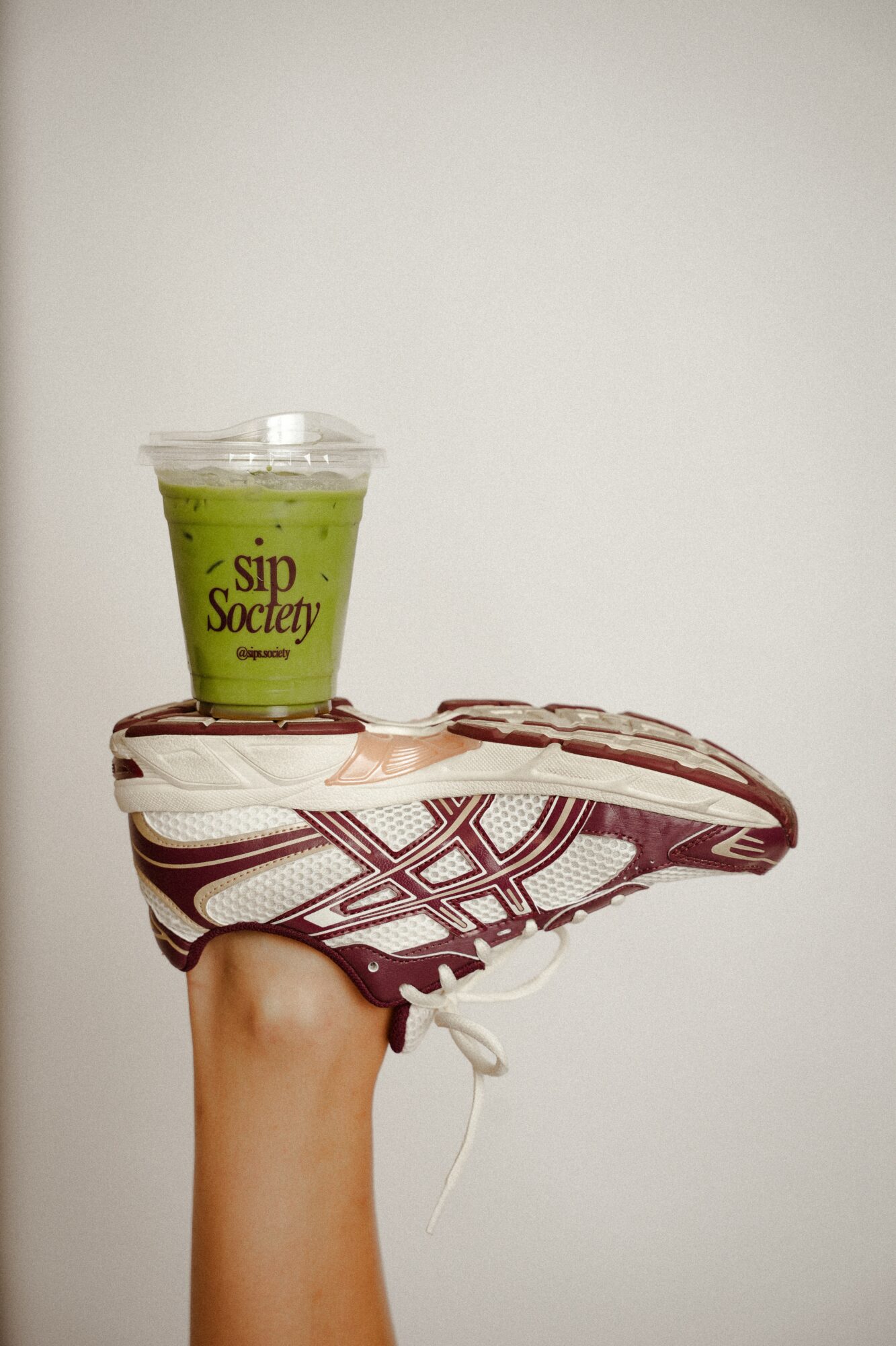 A hand holding a sneaker with a green drink on top, against a plain background.