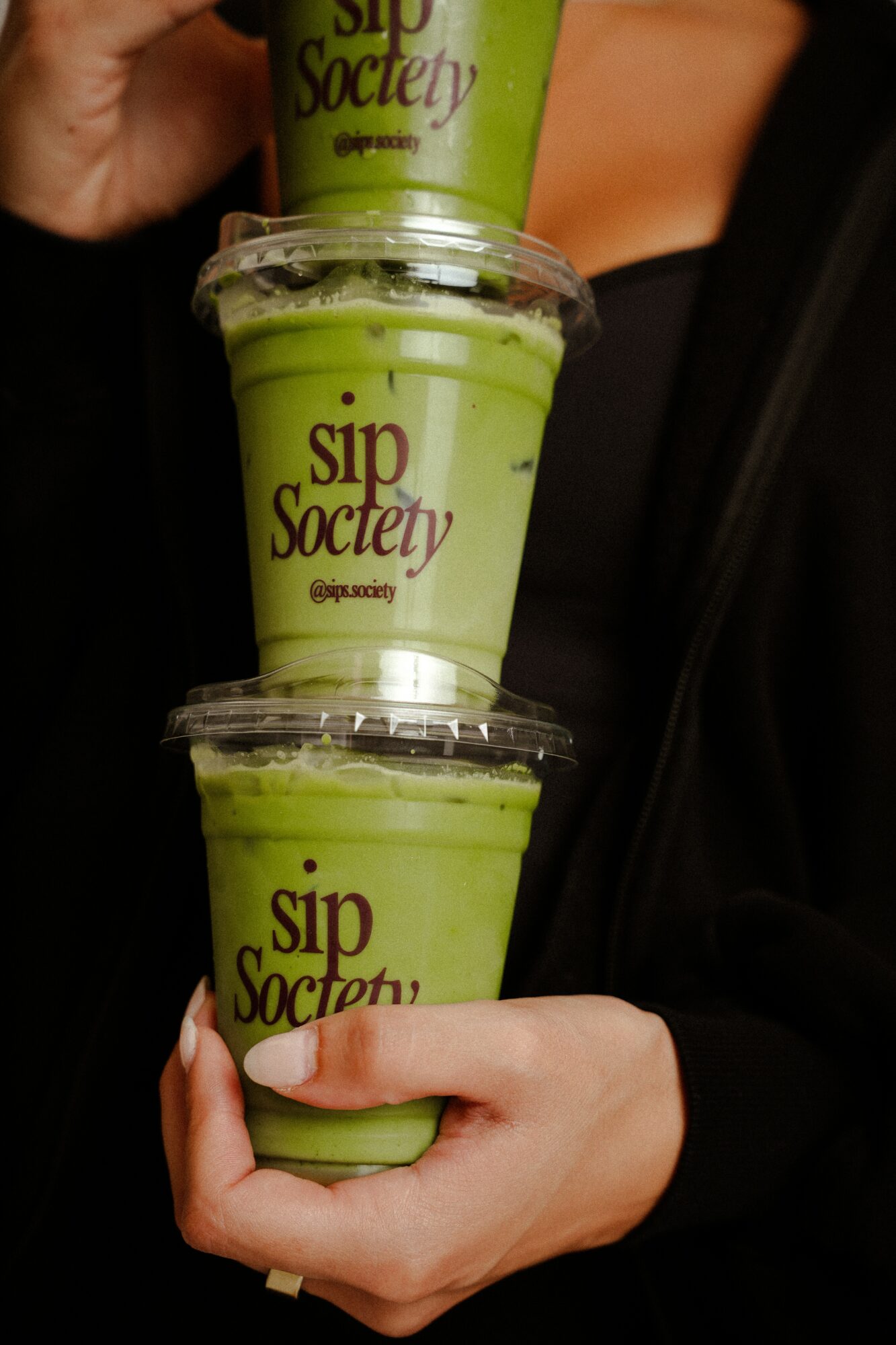 Person holding three green drinks in clear cups stacked vertically, with a hand visible at the bottom and a blurred background.