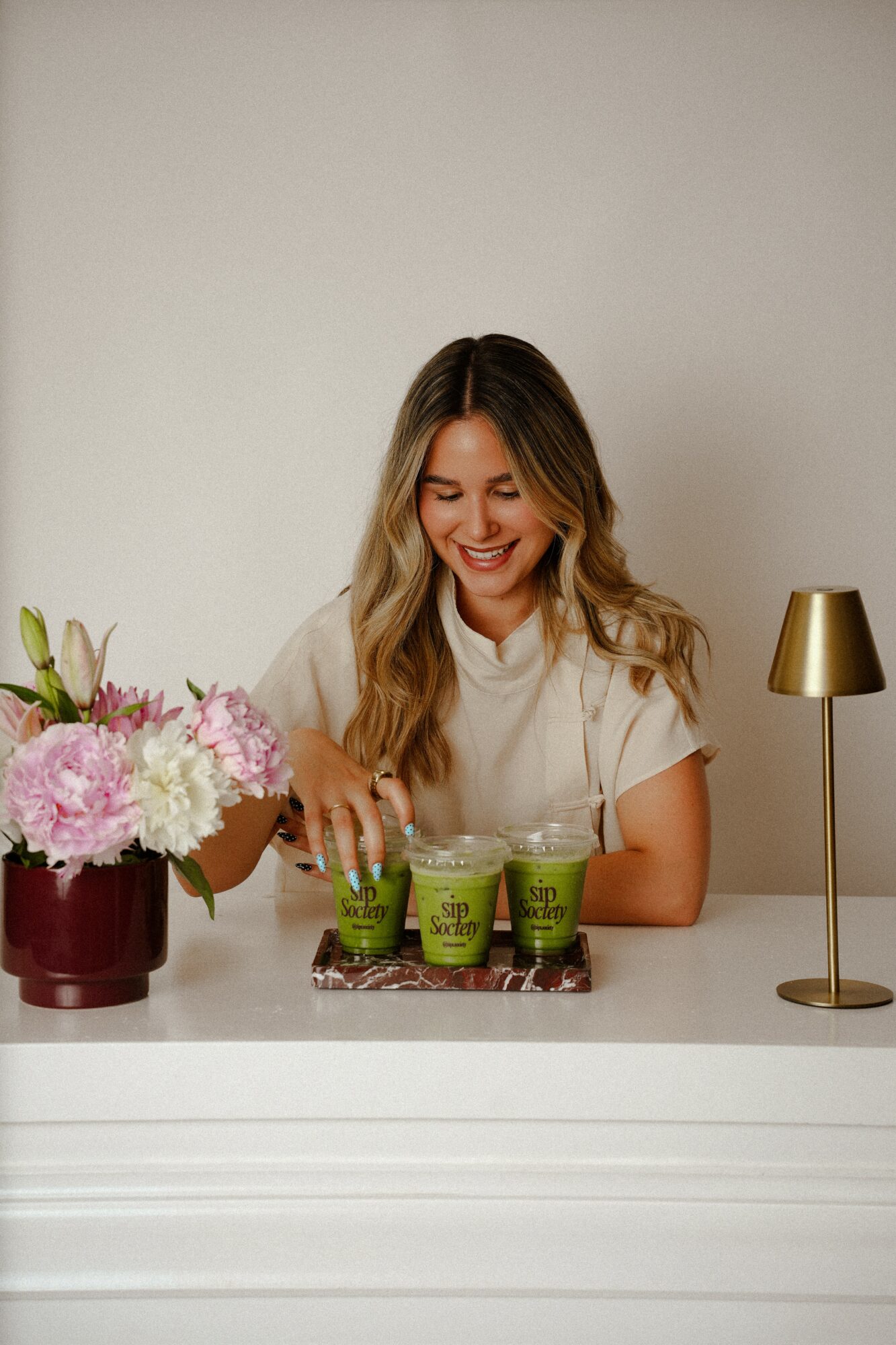 Woman with long hair smiling at a table with three green drinks, flowers, and a gold lamp.