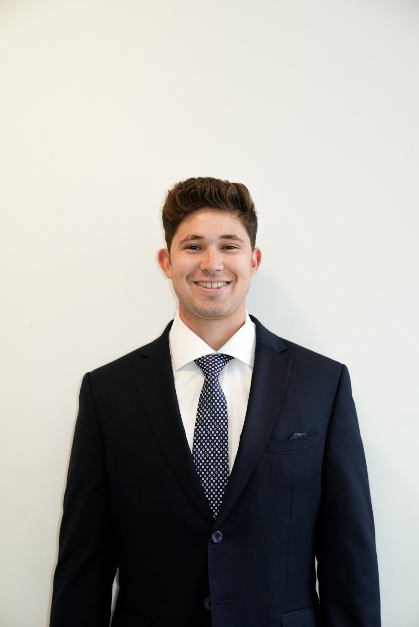 Young man in a dark suit, white shirt, and tie, smiling against a plain white background.