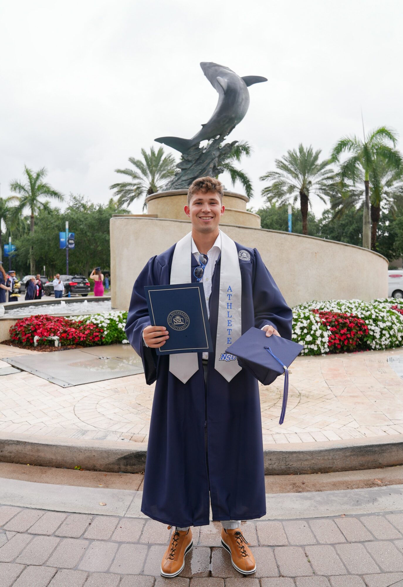 Young man in graduation gown holding diploma and folder, standing outdoors in front of a dolphin statue and palm trees.