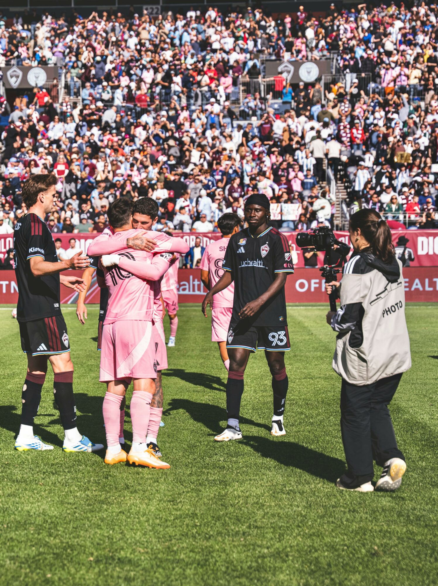 Group of soccer players and a person with a camera on a field, with a large crowd in the background.