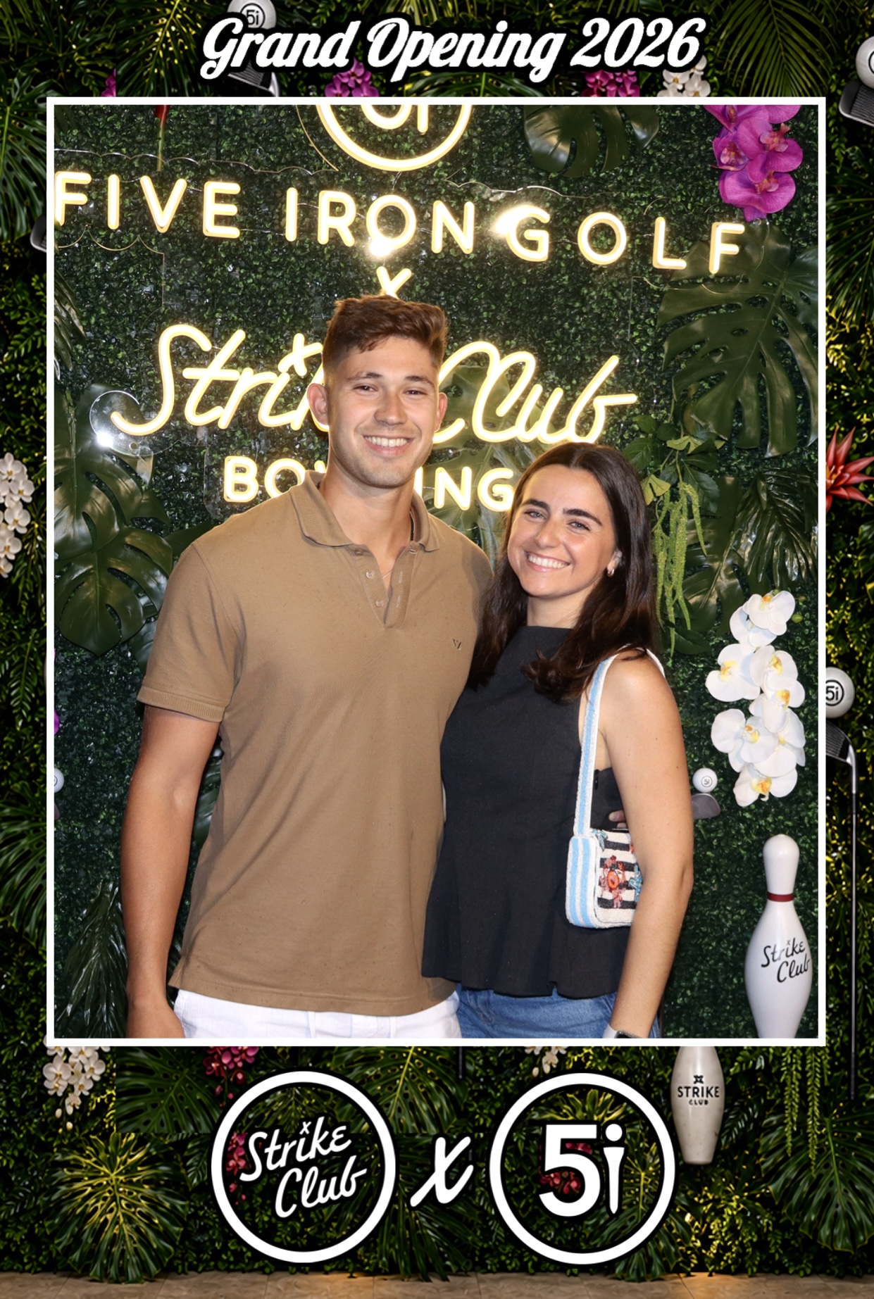 Two people smiling in front of a sign for Five Iron Golf and Strike Club, with floral decorations and a bowling pin.