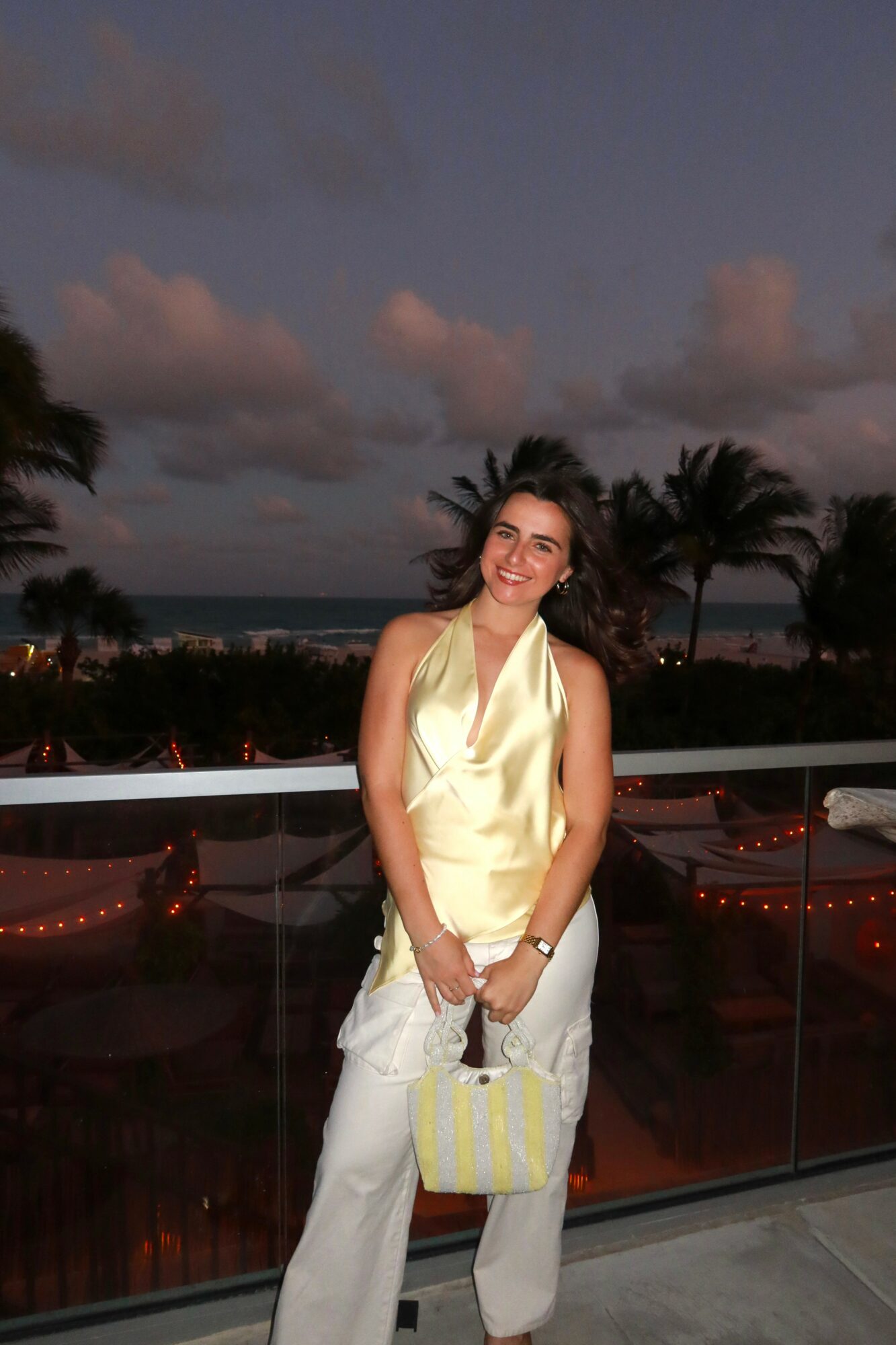 Young woman smiling outdoors at sunset, wearing a yellow top and white pants, holding a yellow bag, with palm trees in background.