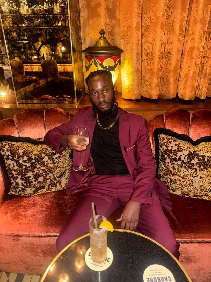 Man in maroon suit sitting on a velvet sofa with pillows, holding a cocktail, in a warmly lit room with curtains and a lamp.