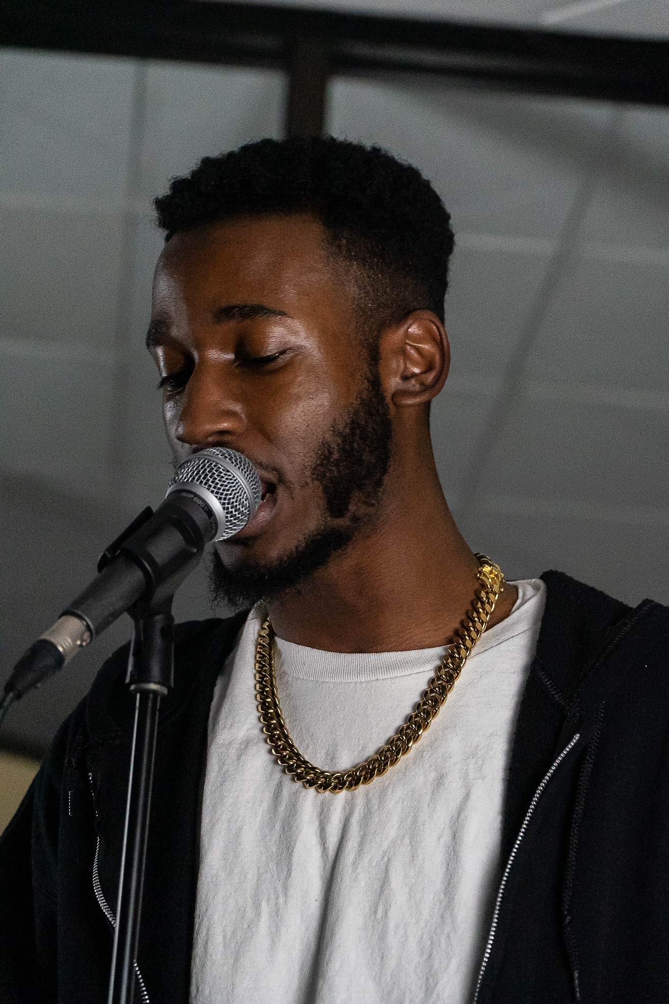 Young man singing into a microphone, wearing a white shirt, black jacket, and gold chain, with a ceiling background.
