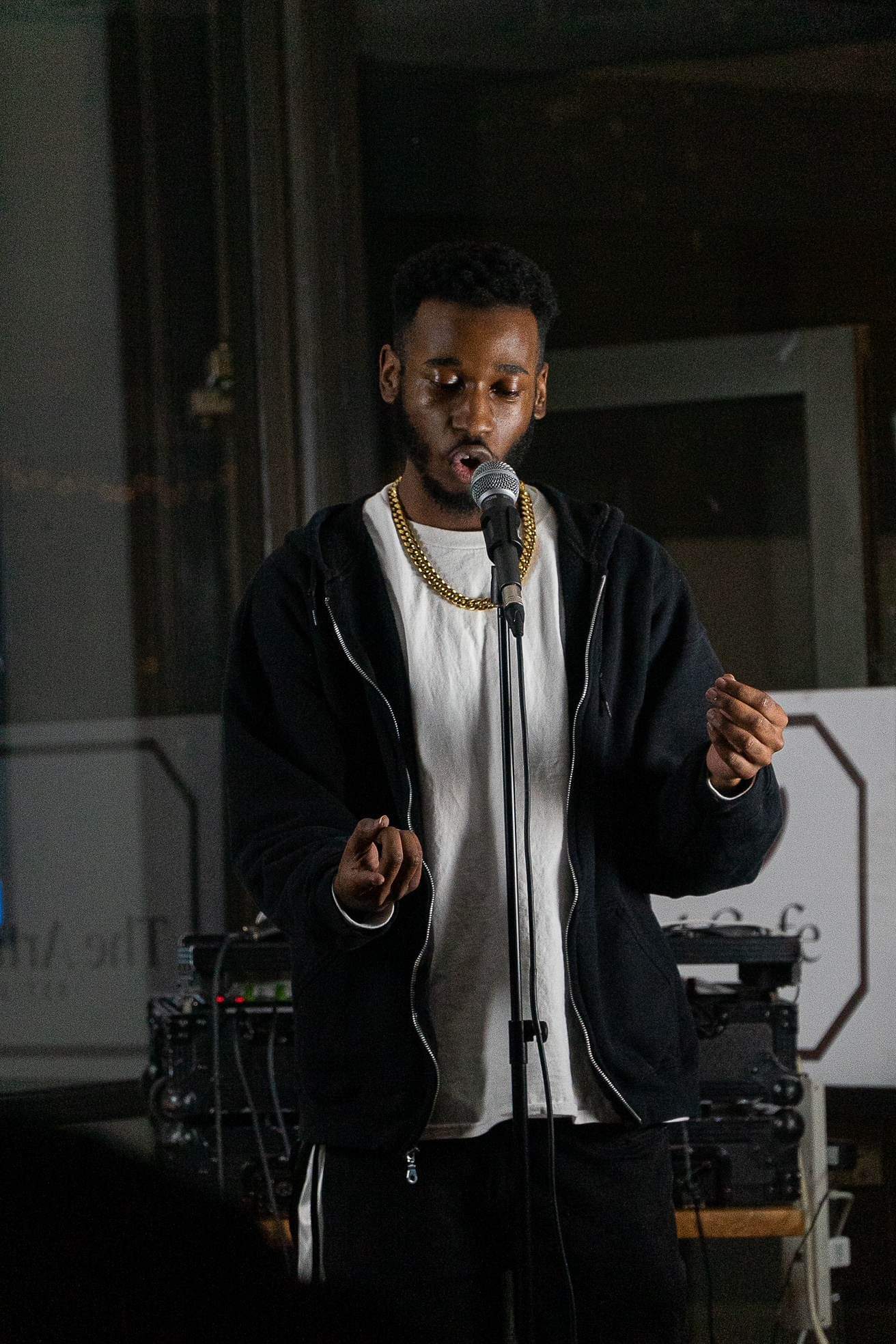 Young man speaking into a microphone, wearing a black hoodie and gold chain, in a dimly lit indoor setting.