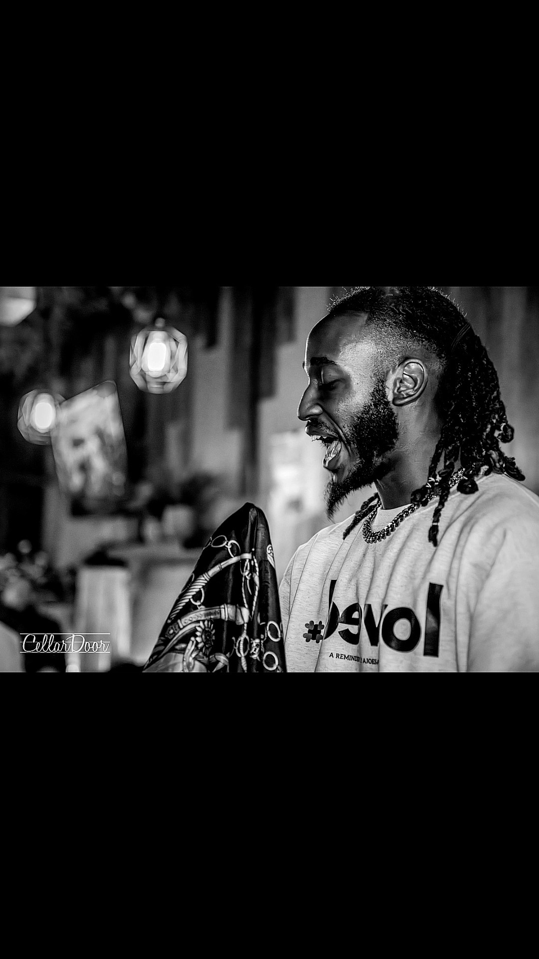 Person with braided hair smiling, wearing a shirt with text, in a room with lights and objects in the background.