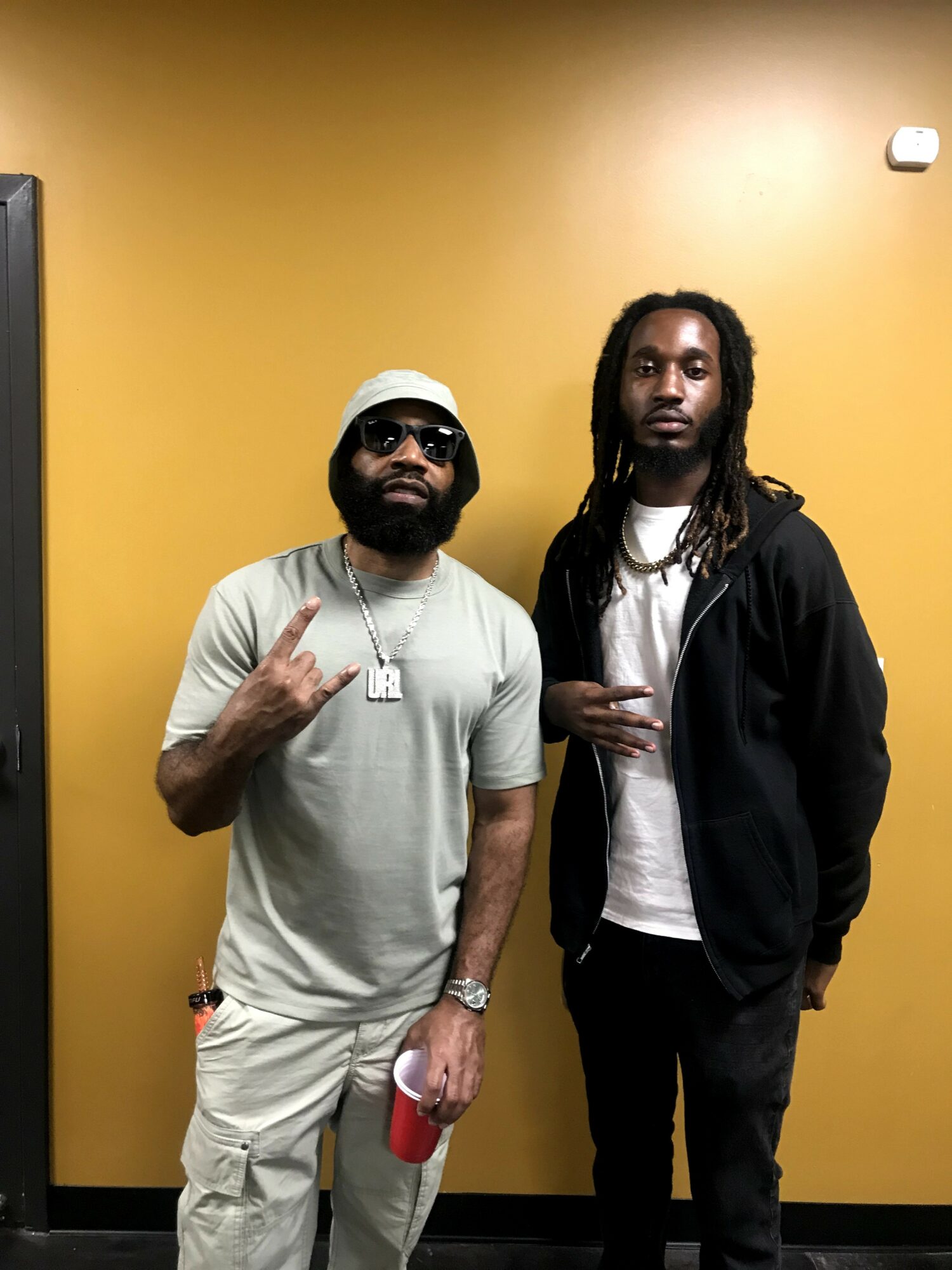 Two men standing against a yellow wall, one wearing sunglasses and a cap, the other with long dreadlocks, both casually dressed.